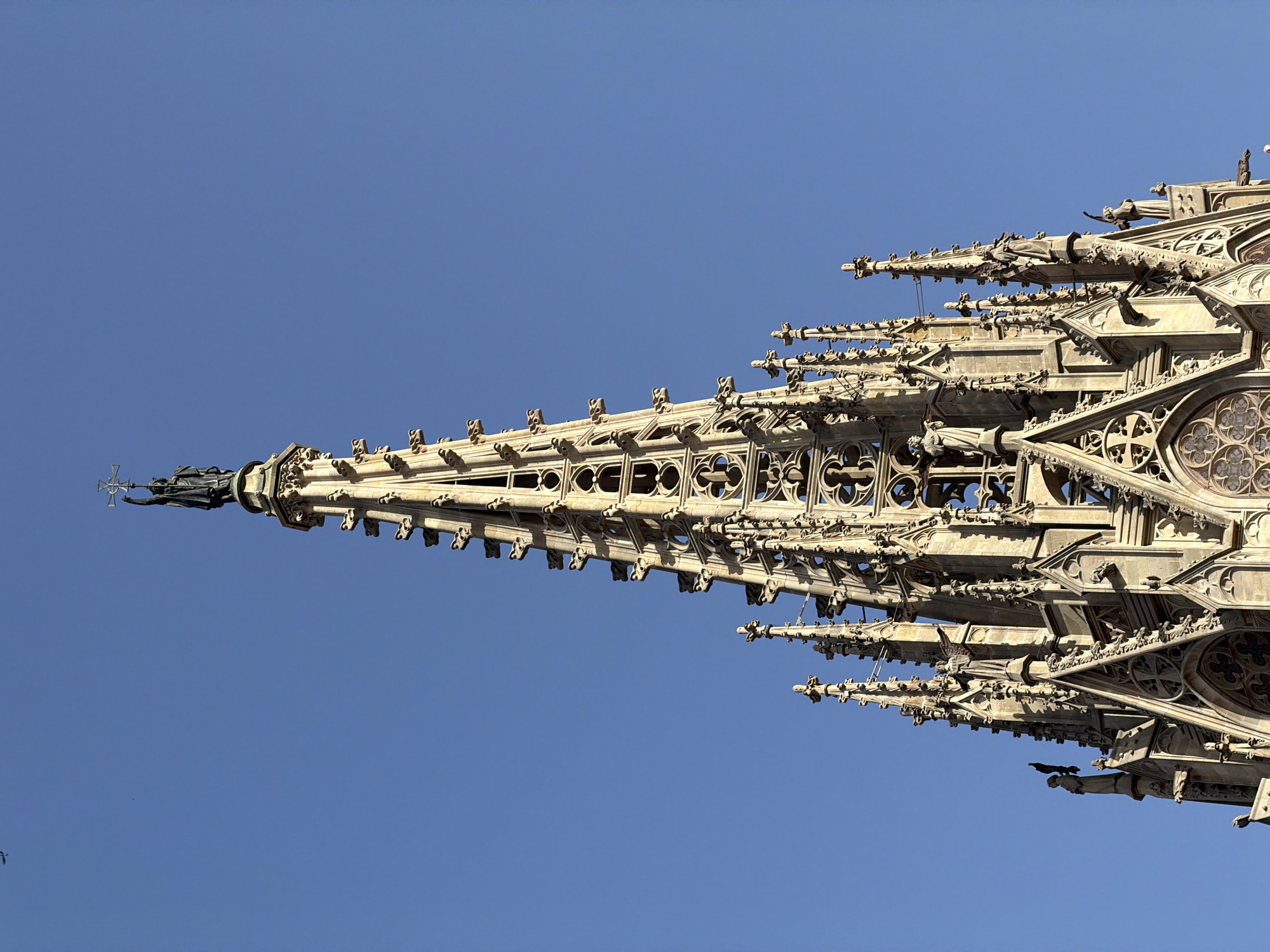This is a close-up, low-angle exterior shot of the spires of the Barcelona Cathedral, a Gothic Roman Catholic cathedral located in Barcelona, Spain. The detailed stonework of the spires, adorned with intricate tracery, gargoyles, and statues, is prominently featured against a clear, bright blue sky. At the very apex of the central spire, a dark statue of a figure holding a cross stands silhouetted. The sun is illuminating the right side of the spires, casting subtle shadows. There are no people visible in the image.