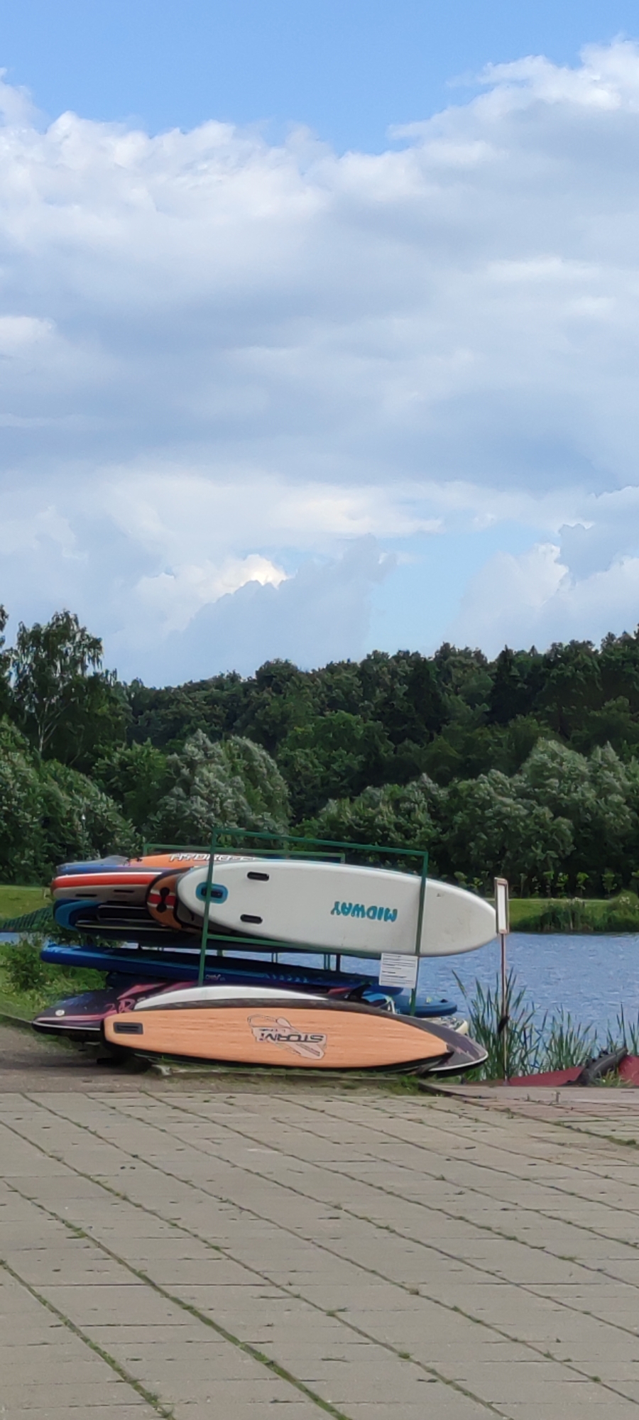 An outdoor scene in Rosinka, Russia, features a rack of stand-up paddleboards (SUPs) on a paved area near a body of water. The SUPs are stacked on a metal rack, with the topmost board being white with the blue text "MIDWAY" printed sideways. Below it are several other SUPs in various colors including orange, red, blue, and black with a multi-colored design. In the background, a dense line of green trees forms a natural backdrop, and a calm body of water is visible to the right. The sky above is a light blue with scattered white clouds. The foreground consists of a grey paved surface with visible grout lines. No people are present in the image.