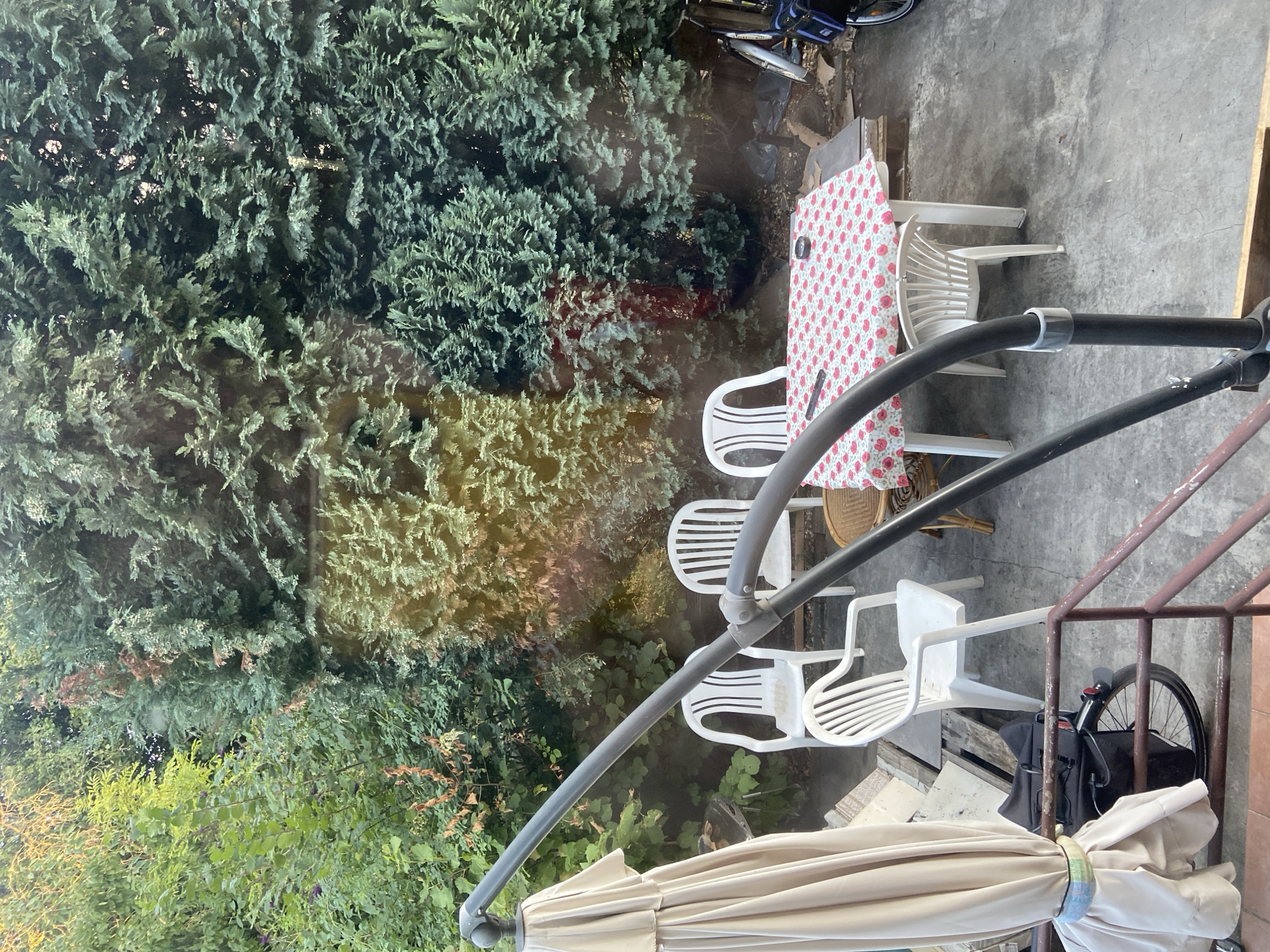 This is an outdoor scene of a small patio area. In the foreground, a portion of a closed beige umbrella and a dark grey metal support pole are visible. Beyond this, a patio table is covered with a red and white floral tablecloth, set with what appear to be four white plastic chairs and one wicker chair. A bicycle with a black bag is parked to the right of the table. The background is dominated by dense green evergreen trees, suggesting a private garden or yard. The scene appears to be in a residential area.