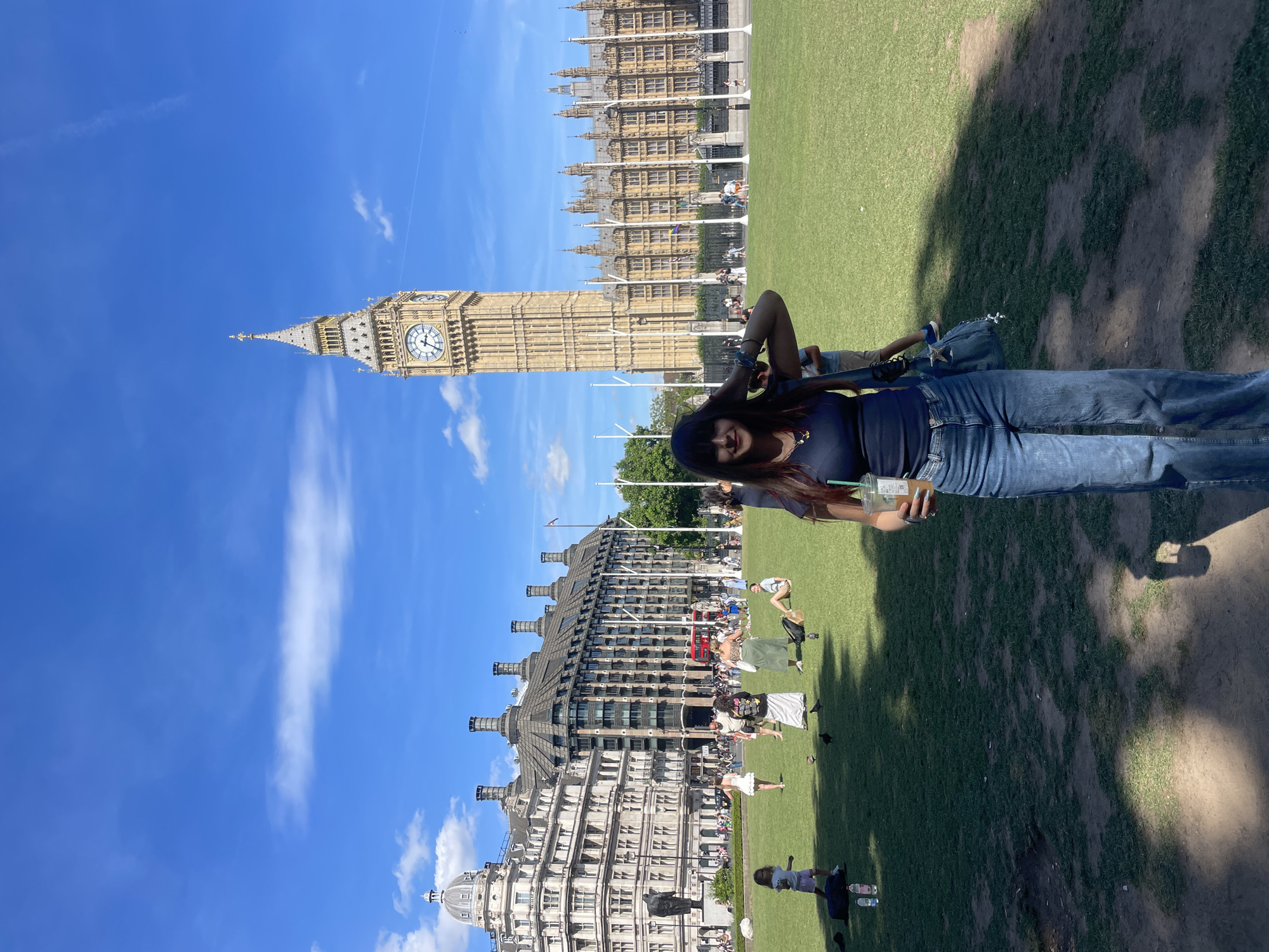 A woman stands in a park in front of the Elizabeth Tower (commonly known as Big Ben) and the Houses of Parliament in London, United Kingdom. The woman is wearing a dark blue tank top and high-waisted, light-wash jeans. She is holding a cup with a straw in her right hand and has her left hand on her head, touching her hair. A blue purse with a star charm hangs from her left shoulder. She is smiling at the camera.
In the background, people are scattered across a large grassy area. Some are sitting on the grass, while others are walking or standing. Children are also present in the park. The iconic architecture of the Houses of Parliament and the Elizabeth Tower dominate the background, with a clear blue sky above. The sun casts shadows on the grass, indicating it is a bright, sunny day.