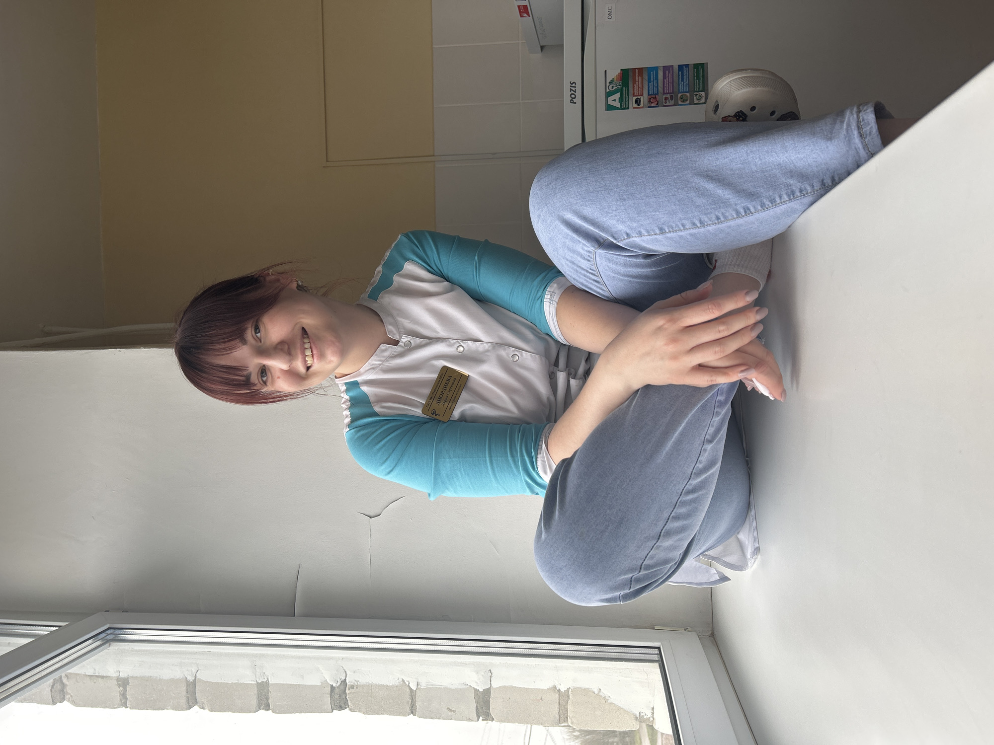 A woman in a medical uniform sits cross-legged on a white surface by a window. The uniform has white fabric with light blue sleeves. She has brown hair and is smiling. Her name tag is in Russian. The window looks out onto a brick exterior. There is a refrigerator labeled "POZIS" in the background. The location is Фоки, Russia.