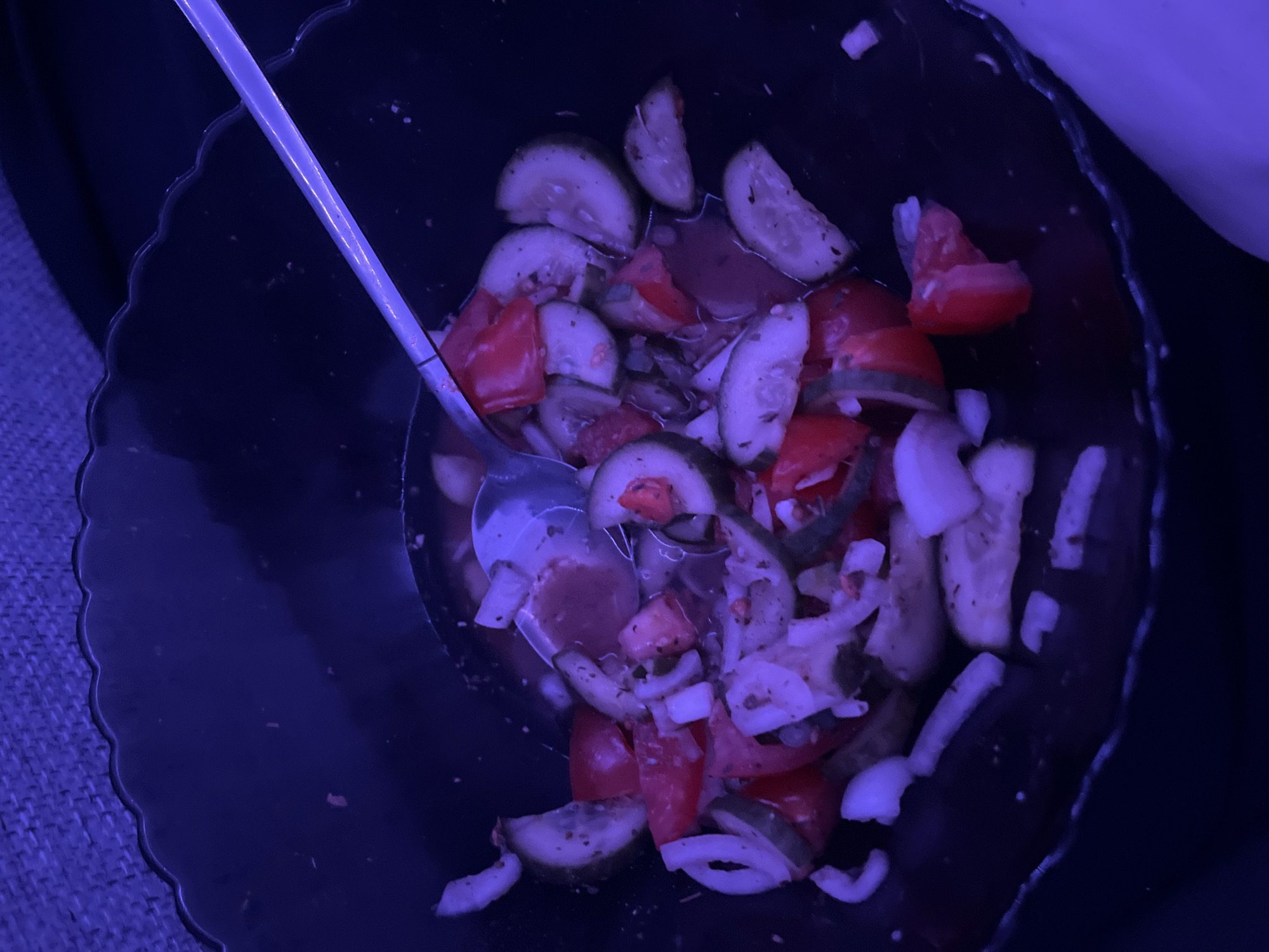 The image shows a bowl of salad with a spoon. The salad consists of sliced cucumbers, tomatoes, and onions in a dark liquid. The bowl appears to be dark-colored and has a scalloped edge. The background is dark and suggests the bowl is placed on a surface, potentially a cushion or fabric. There is no indication of the location being Saint Petersburg, Russia, or any people present in the image.