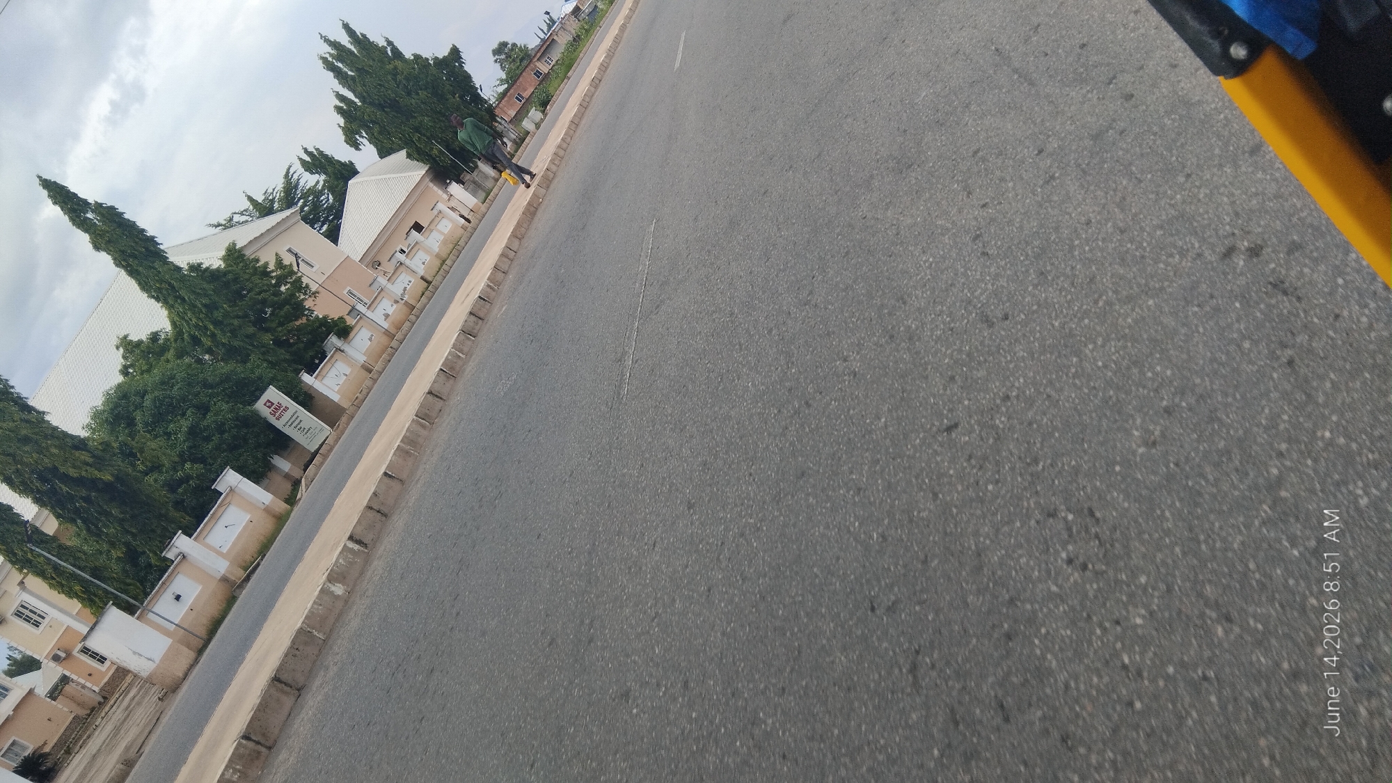 A person in a green shirt and dark pants walks on a paved sidewalk next to a road in Jalingo, Nigeria. They are carrying a yellow bag. In the background, there is a building with a white roof and beige walls, surrounded by tall, dark green trees. A sign with the words "BANLE SUITES" is visible near the building. The sky is cloudy. The scene is captured on June 14, 2026, at 8:51 AM.