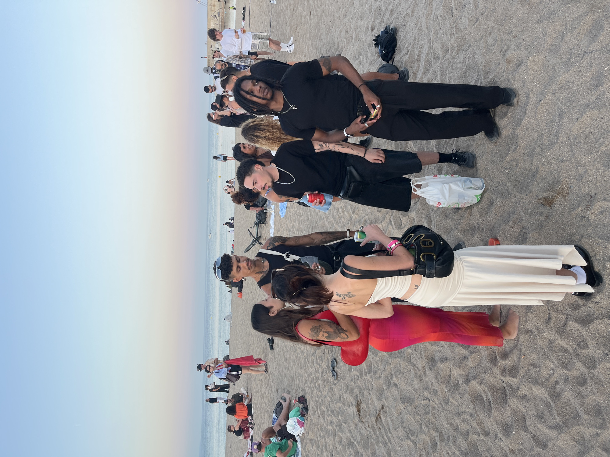 This image captures a late afternoon or early evening scene on a sandy beach in Sant Adrià de Besòs, Spain, overlooking the calm Mediterranean Sea. The sky transitions from a light blue overhead to a pale pink-orange hue near the horizon. A concrete pier or breakwater is visible on the far right.
In the foreground, a group of four individuals is engaged in conversation. From left to right:
1. A woman with dark hair and arm tattoos wears a form-fitting, sleeveless dress that gradients from dark pink to orange. She is barefoot and holds a red heart-shaped bag.
2. A woman with dark hair tied back, revealing a tattoo on her upper back, wears a long, off-white, draped sleeveless dress. She wears white socks with black shoes and carries a black shoulder bag, holding a green can.
3. A man with dark skin and dreadlocks wears a black short-sleeved t-shirt, black pants, and dark shoes. He has arm tattoos, a silver chain with a cross pendant, and rings on his fingers, holding a small yellow object. A black backpack lies on the sand behind him.
4. A man with short dark hair and a mustache wears a black t-shirt, silver chain, black shorts, and dark sneakers. He carries a white plastic shopping bag and holds a red can, with a black fanny pack worn across his chest.
Behind this group, other individuals are scattered across the sand. A man in a black tank top, black pants, and sunglasses pushed onto his head stands near a bicycle. Further back, people are walking, standing, or sitting on towels on the sand, dressed in casual beach attire. A man holding a red towel stands closer to the water's edge in the mid-left. The sand is light brown, marked with footprints.