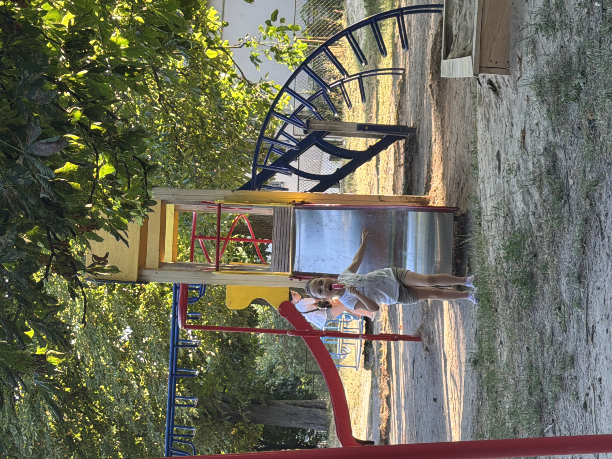 In this image, a young girl stands on a playground slide, holding a pink popsicle. She wears a white t-shirt and grey shorts, with white sneakers on her feet. Her hair is pulled back, and she has a playful expression with her tongue sticking out. Another person, partially visible, is seated on the slide behind her. The playground features a metal slide, a blue climbing structure, and red railings. The surrounding area is sandy with patches of grass and trees with green leaves, suggesting an outdoor setting on a sunny day. The light suggests it is late afternoon. The location is identified as Kamianka-Buzka, Ukraine.