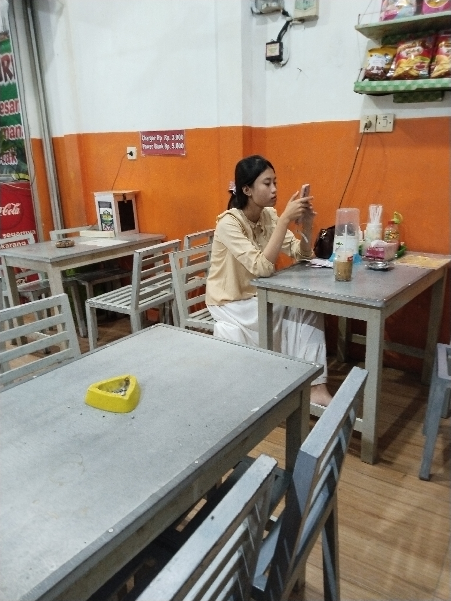 A young woman is sitting at a table in what appears to be a casual dining establishment. She is wearing a light-colored long-sleeved shirt and white pants and is looking at her phone. On the table in front of her are a glass with a beverage, a container of what looks like napkins, and a small bottle. Behind her is an orange wall with a sign indicating "Charger Rp. 3.000 Power Bank Rp. 5.000" and a white charging station. The establishment has wooden floors and tables and chairs made of a light-colored material. Another table with a yellow ashtray is in the foreground. This location is identified as Prigi, Indonesia.