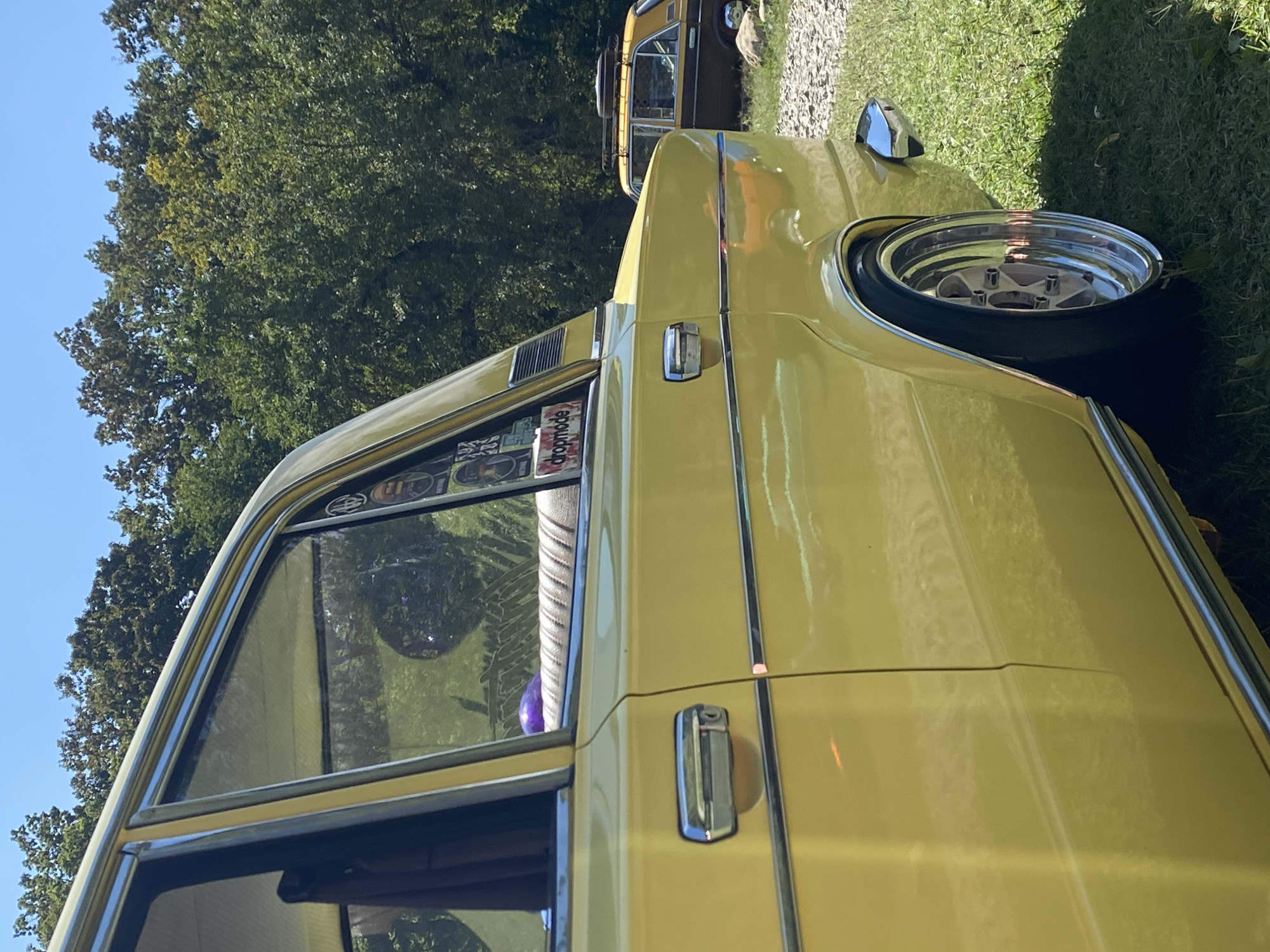 A yellow sedan is parked on grass, with trees and a clear blue sky in the background. The car is viewed from the side, showcasing its chrome door handle and a custom wheel. Another car, a brown sedan, is visible in the distance. The image appears to have been taken during the daytime, with sunlight reflecting off the car's surface. The location is identified as Bezymyannoe, Russia.