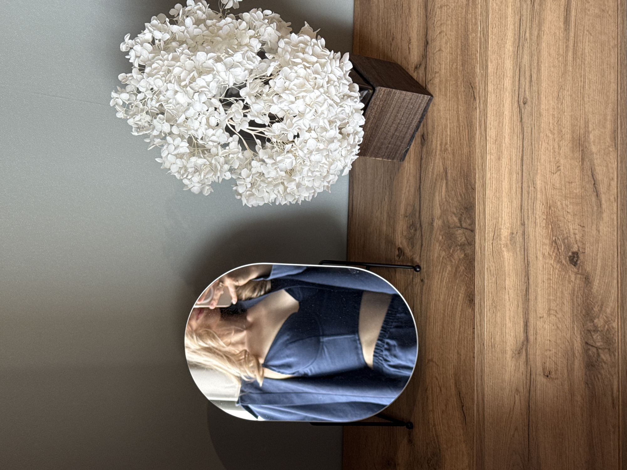 The image presents an overhead view of a domestic interior. A wall painted a light grey is visible on the left side, juxtaposed with a section of wood flooring on the right. A bouquet of white flowers is placed near a dark, wooden rectangular vase or planter. Directly below the flowers and planter is a small, oval mirror resting on a black metal stand. The mirror reflects a person with blonde hair wearing a blue top. The reflection also captures a hand holding a cell phone. There's no indication that this image was taken in the city of Chaikovsky, Russia.