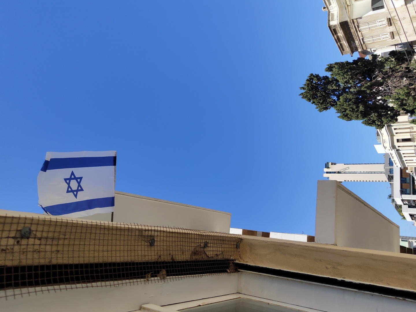 The image presents a bright, clear blue sky over an urban landscape, identified as Tel-Aviv, Israel, due to the prominent national flag. In the lower left foreground, a weathered wooden structure covered with a wire mesh, possibly a protective screen over a window or ventilation, is visible. Below this, part of a white building or balcony structure is also seen.
Above the mesh structure and slightly to the left, a white Israeli flag, emblazoned with two horizontal blue stripes and a central blue Star of David, waves gently against the vibrant sky. Its presence is a strong focal point.
The right side of the frame is bordered by a mix of urban architecture. An older-style building with light-colored walls and some ornate details occupies the upper right. Below it, a large, dark green tree, possibly a cypress, stands out against the sky. Further in the background, a tall, modern high-rise building with a distinct grid pattern is visible, contributing to the city skyline.
The bright, clear blue sky and strong daylight indicate a sunny day. No specific human activity is depicted. There is no discernible text in the image. The scene conveys a sense of peaceful urban life under national display.