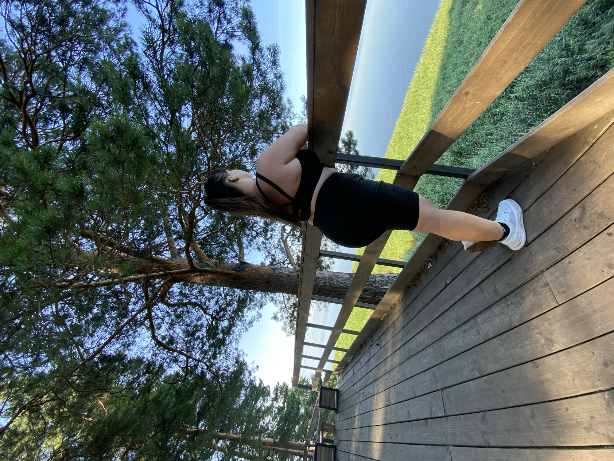 The image depicts a woman standing on a wooden deck overlooking a body of water. She is wearing black athletic wear and white sneakers. The deck is elevated, with a wooden railing. Pine trees are visible in the foreground and background. The sky is clear and blue. The scene suggests a natural outdoor setting, possibly a park or a viewpoint. The specific location in Votkinsk, Russia, is not discernible from the image itself, but it is likely a recreational area with a view of the water.