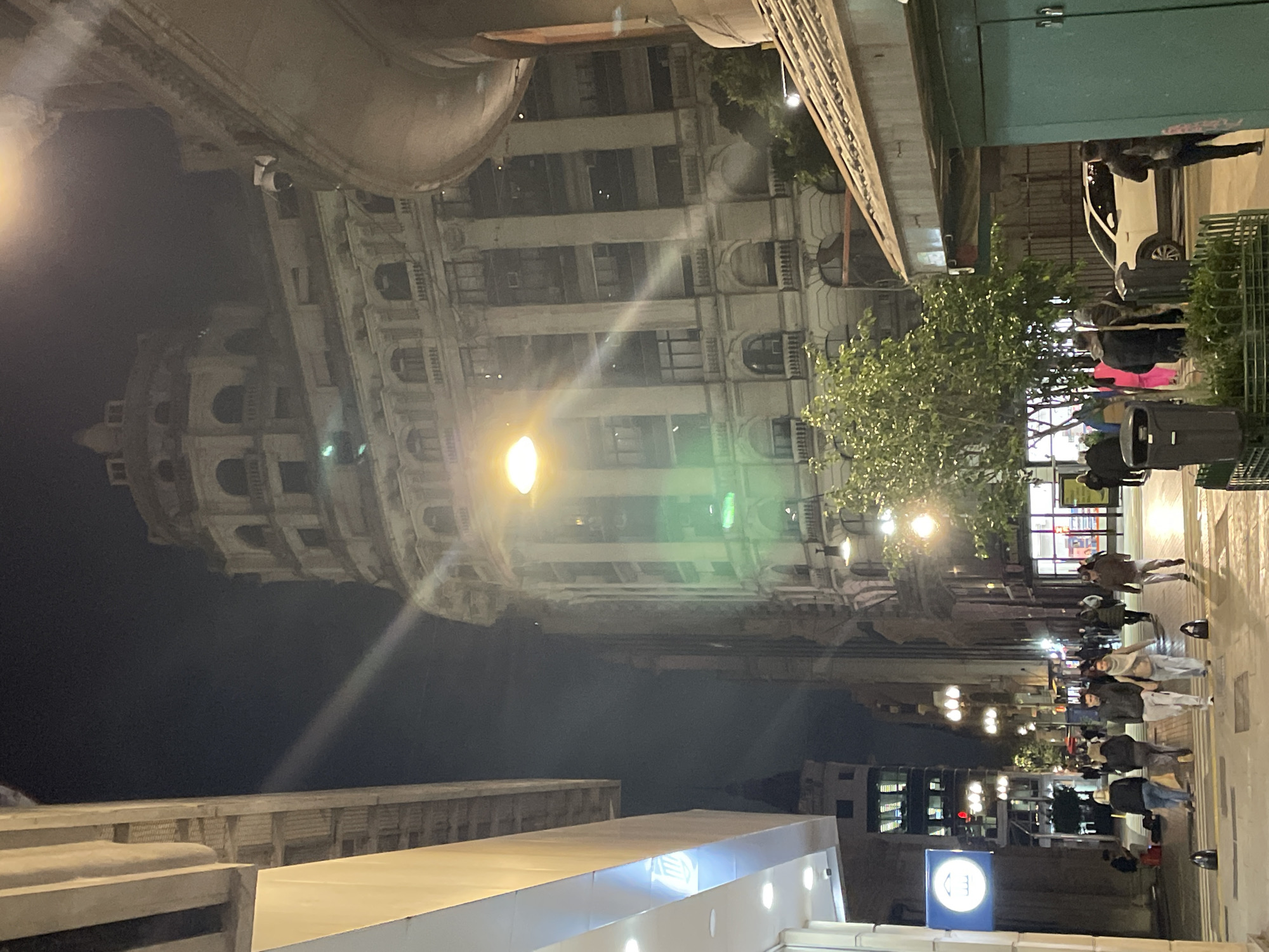 An eye-level, outdoor nighttime shot shows the dome and exterior of the Galerías Pacífico in Buenos Aires, Argentina, with a sidewalk and a crowd of people moving along the sidewalk in the foreground. The sky is dark, while the building's facade and the sidewalk are illuminated by streetlights. The building is tall and ornate, with multiple windows and architectural details visible. The people on the sidewalk are walking in both directions; their actions and interactions are typical of a busy city street at night. Some people are in pairs. A white car is parked on the street. The street and sidewalk appear to be well-maintained.