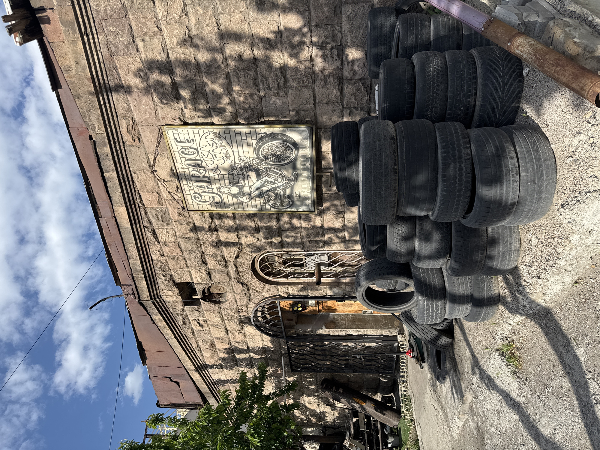 The image depicts the exterior of a building in Yerevan, Armenia. The building is constructed of stone and features an arched doorway with metalwork. A sign with the word "GARAGE" and a motorcycle illustration is hung on the building's facade. Stacks of used tires are piled to the right of the entrance. The foreground shows a portion of a tree and various objects, suggesting the scene is a working garage environment. The sky with clouds is visible in the upper left corner. The light suggests it is a sunny day.
