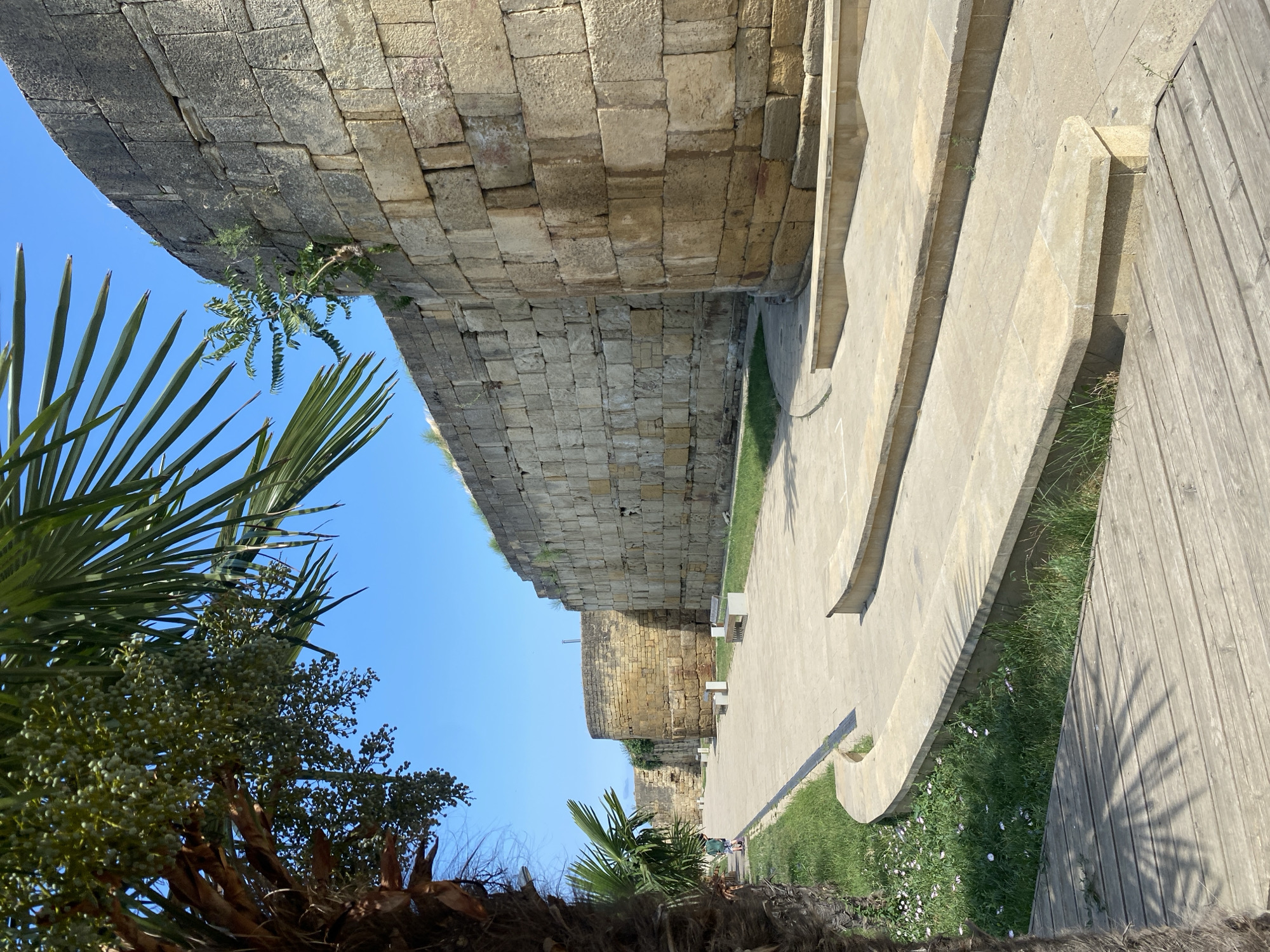 The image depicts a section of the ancient city walls in Derbent, Russia, a UNESCO World Heritage site. The stone walls, constructed from large, weathered blocks of yellowish-tan stone, curve around a paved walkway. The wall on the right is notably curved and appears to be a defensive tower or bastion. In the foreground, a wooden boardwalk runs diagonally, leading the eye towards the path and the imposing wall. Beyond the paved walkway, there is a strip of green grass with small white flowers, and further back, a younger stone wall with a rounded section is visible. Palm trees are present on the left side of the image, with their fronds casting shadows on the boardwalk and the grassy area. The sky is a clear, bright blue. A lone figure, likely a tourist, is walking on the paved path in the distance, heading towards the curved section of the wall. Several modern-looking stone benches are placed along the walkway. The scene suggests a sunny day, with the architecture indicative of a historical fortress or citadel.