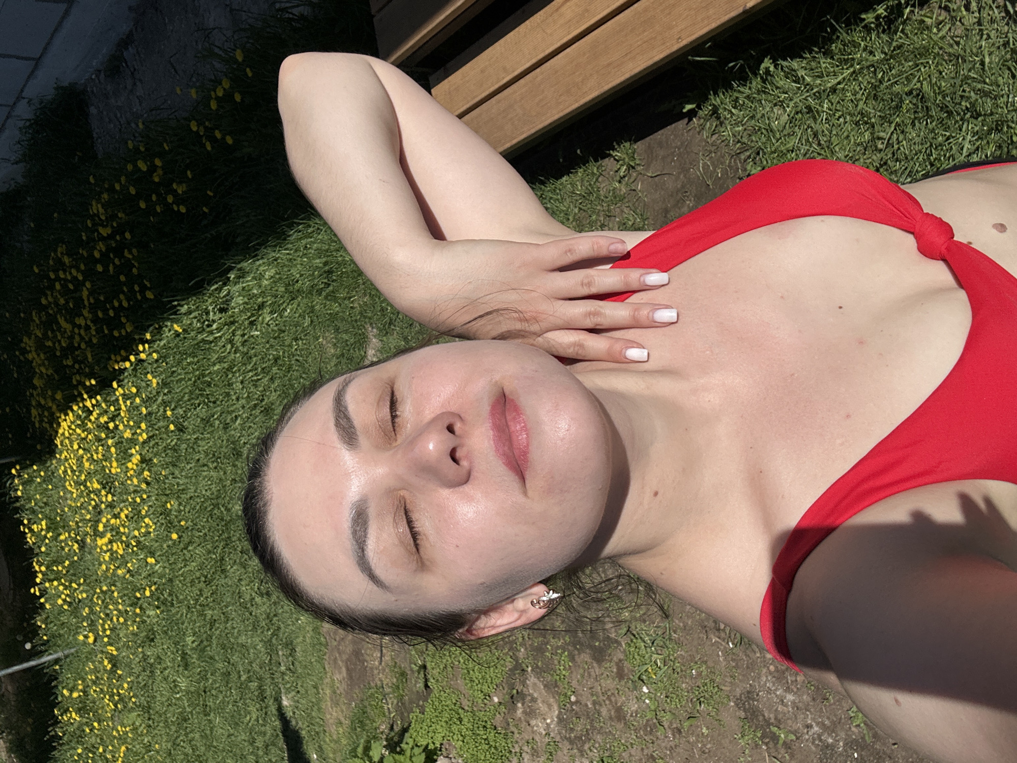 A woman is lying on grass in Chaikovsky, Russia. She is wearing a red swimsuit. She has fair skin, dark hair, and is lying on her back with her eyes closed. Her left arm is raised, with her hand resting on her collarbone. The background includes grass with yellow flowers. There is a wooden structure partially visible in the upper part of the image.