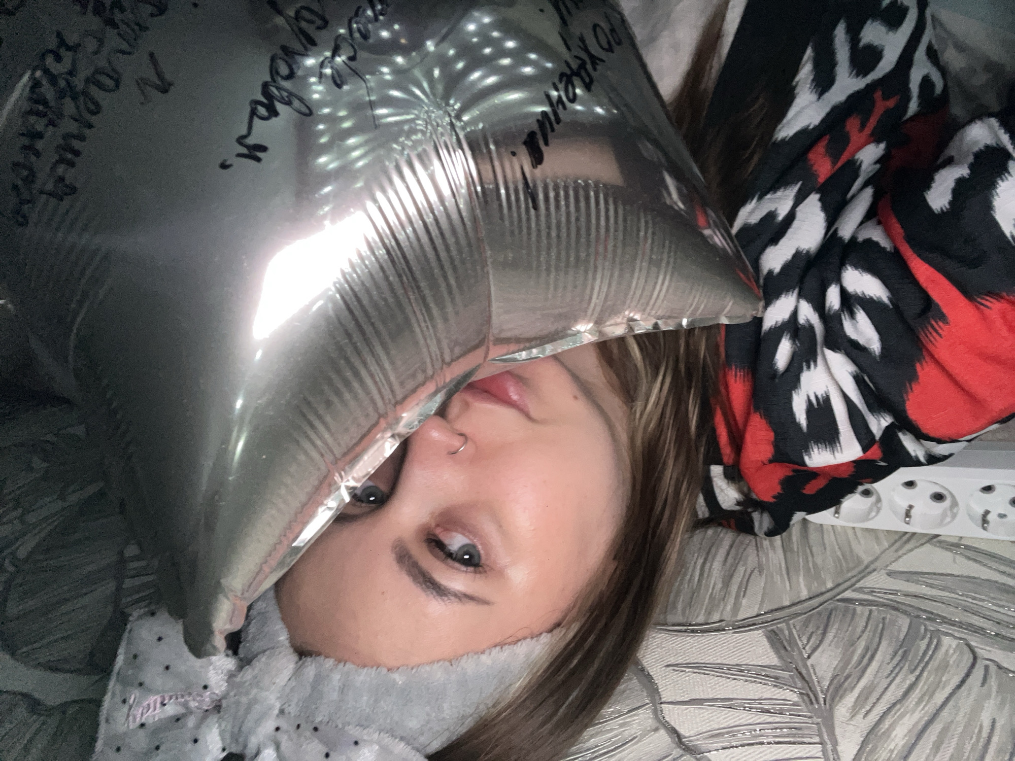 A woman is lying down with a silver helium balloon near her face. The balloon has handwritten words on it in black marker. She has fair skin, grey eyes, and a nose ring. She is wearing a grey, fuzzy headband. Next to her is a patterned blanket in red, black, and white. The woman is on a leaf-patterned blanket on a bed. In the background is a white power strip. The location is Novopokrovskaya, Russia.