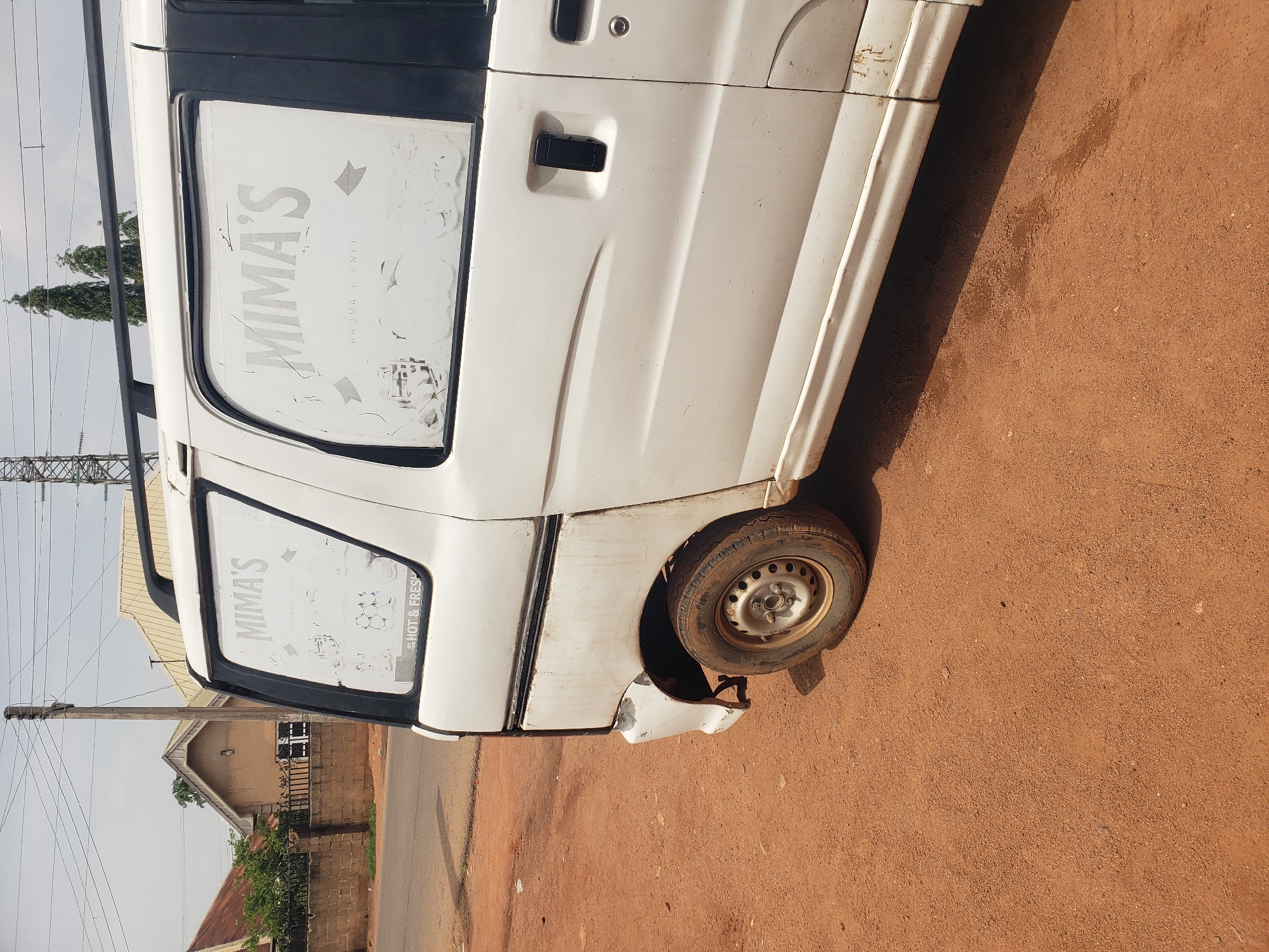 In Bukuru, Nigeria, a white minivan is parked on a dirt road. The vehicle has several decals on its rear windows that appear to read "MIMA'S" and "BROVATE FREE." The minivan shows signs of wear, including scratches and rust on its body. The rear passenger-side wheel is visible, and the tire is mounted on a silver rim. The surrounding environment consists of a paved road to the left, a brick wall, a house with a metal roof, and several trees in the background. Power lines and a utility pole are also present in the sky. The ground is covered with reddish-brown dirt and some sparse vegetation. The sky is overcast.
