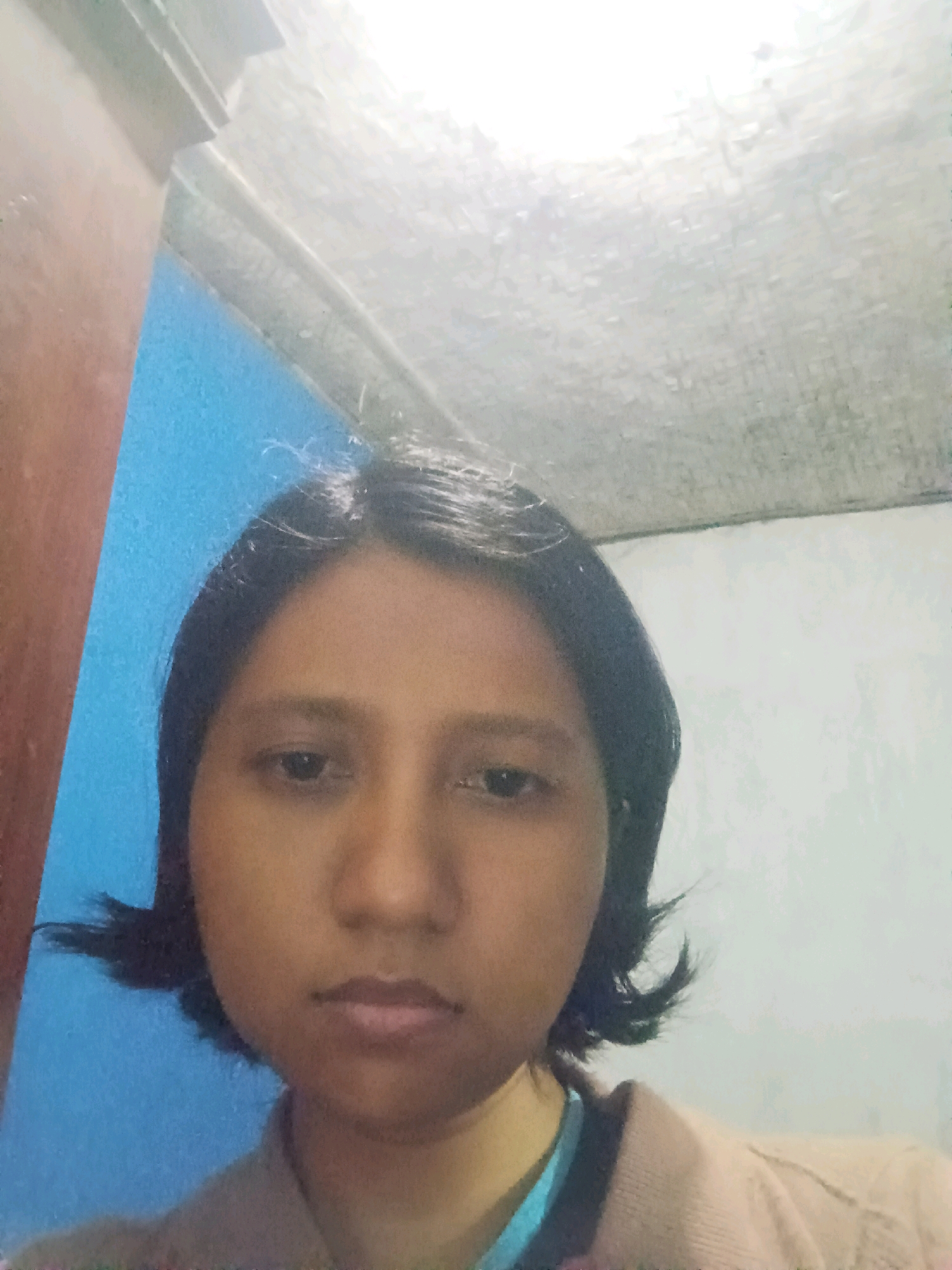 A head and shoulders portrait of a young woman with dark hair and brown eyes is shown. She is looking directly at the camera. The background consists of a blue wall to the left and a white wall to the right. The ceiling is visible at the top of the frame. The woman is wearing a light brown jacket over a blue shirt. The image appears to have been taken indoors.