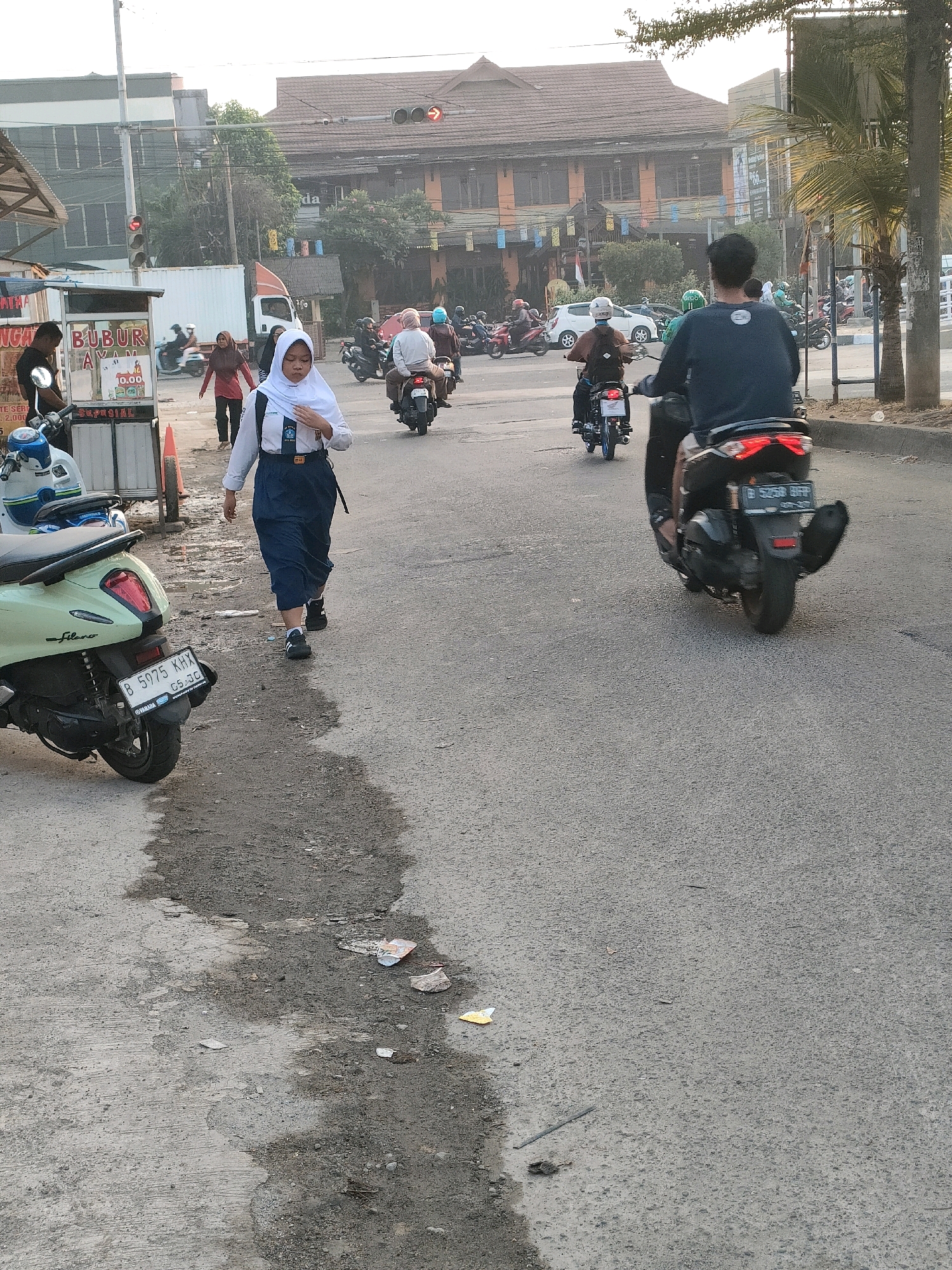 The image depicts a street scene in Bekasi, Indonesia, characterized by a road with some patches of wetness and debris. In the foreground, a light green scooter is parked on the left side, with its license plate reading "B 5975 KHX." A young person wearing a white hijab and a dark blue uniform, possibly a school uniform, walks towards the right side of the frame. Behind this person, another individual in dark clothing rides a black scooter with a license plate that appears to read "B 3059 BHT." In the background, several other motorcycles are visible, some carrying passengers. Traffic lights are present, with one red light illuminated. Buildings line the street, including one with a tiled roof and several stories, and a smaller structure with awnings. The sky is overcast, suggesting it might be daytime with diffused light. The overall atmosphere appears to be that of a busy urban street with ongoing traffic and pedestrian activity.