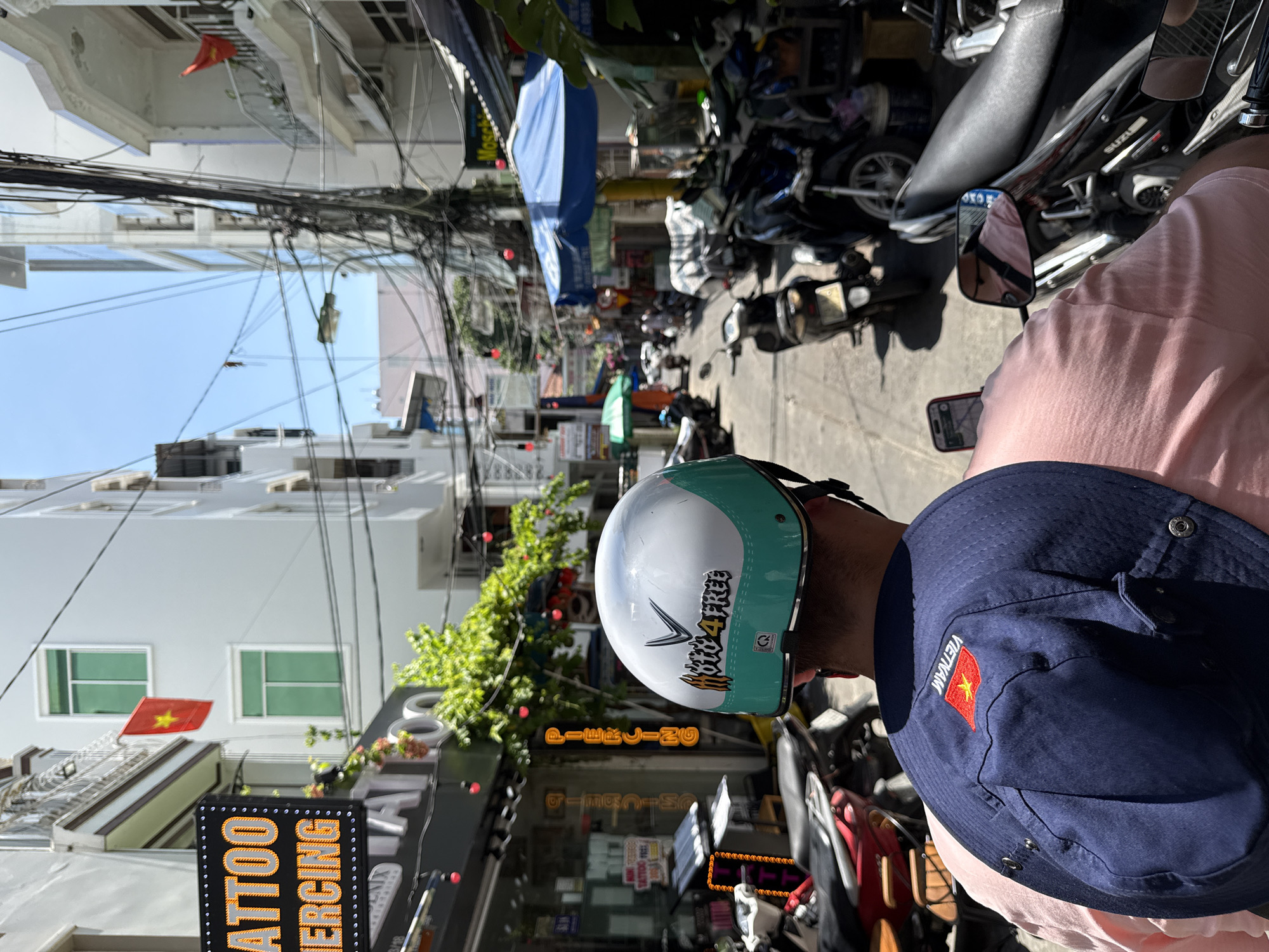 The image depicts a street scene in Nha Trang Ward, Vietnam, captured from the perspective of someone sitting on a motorcycle. The view down the street shows a dense urban environment with buildings, power lines, and parked motorcycles. In the foreground, the back of a person wearing a pink shirt and a blue bucket hat with a Vietnamese flag is visible. They are also wearing a white helmet with green trim and the inscription "foto 4 FREE." To the left, a sign for "TATTOO PIERCING" is prominent, along with a Vietnamese flag displayed on a building. The street is lined with shops and other establishments.