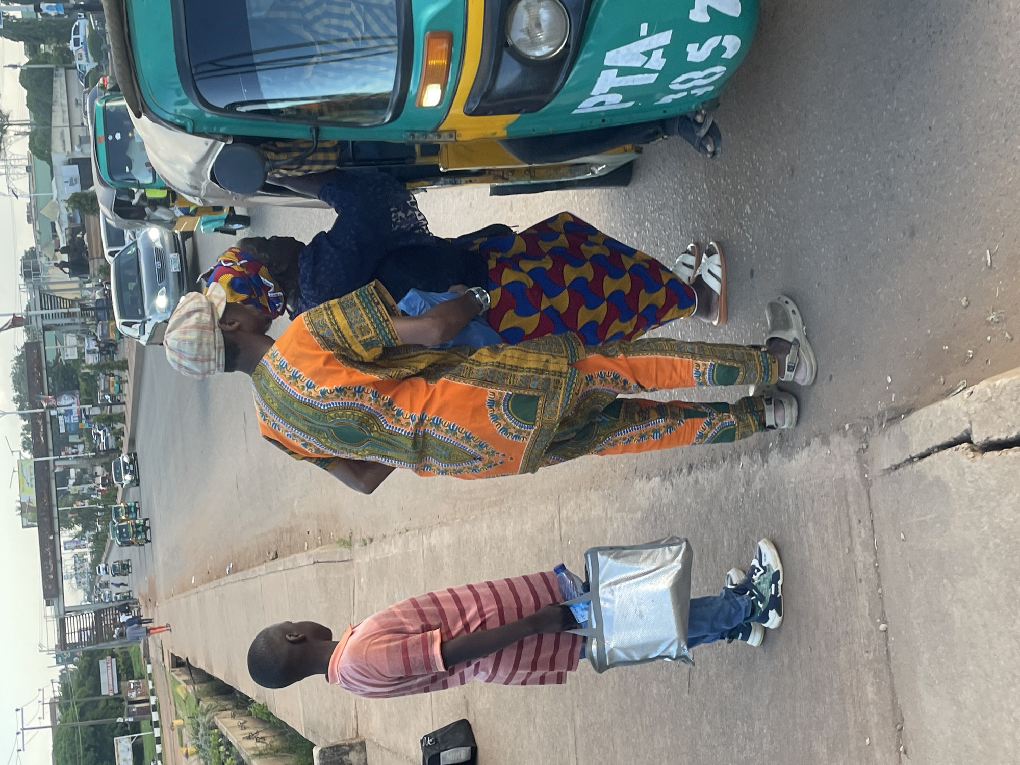 A street scene in Jos, Nigeria, features a young boy in a pink and maroon striped shirt and blue jeans, holding a silver bag. Beside him, a man in a colorful orange and gold patterned outfit and a checkered cap converses with a woman wearing a dark blue lace top and a vibrant, patterned skirt. The group stands near the side of a busy road with several auto-rickshaws, including a green and yellow one with "PTA 1852" visible on its side. Cars and other auto-rickshaws are present in the background, along with a pedestrian bridge and roadside vegetation. The sky is overcast.