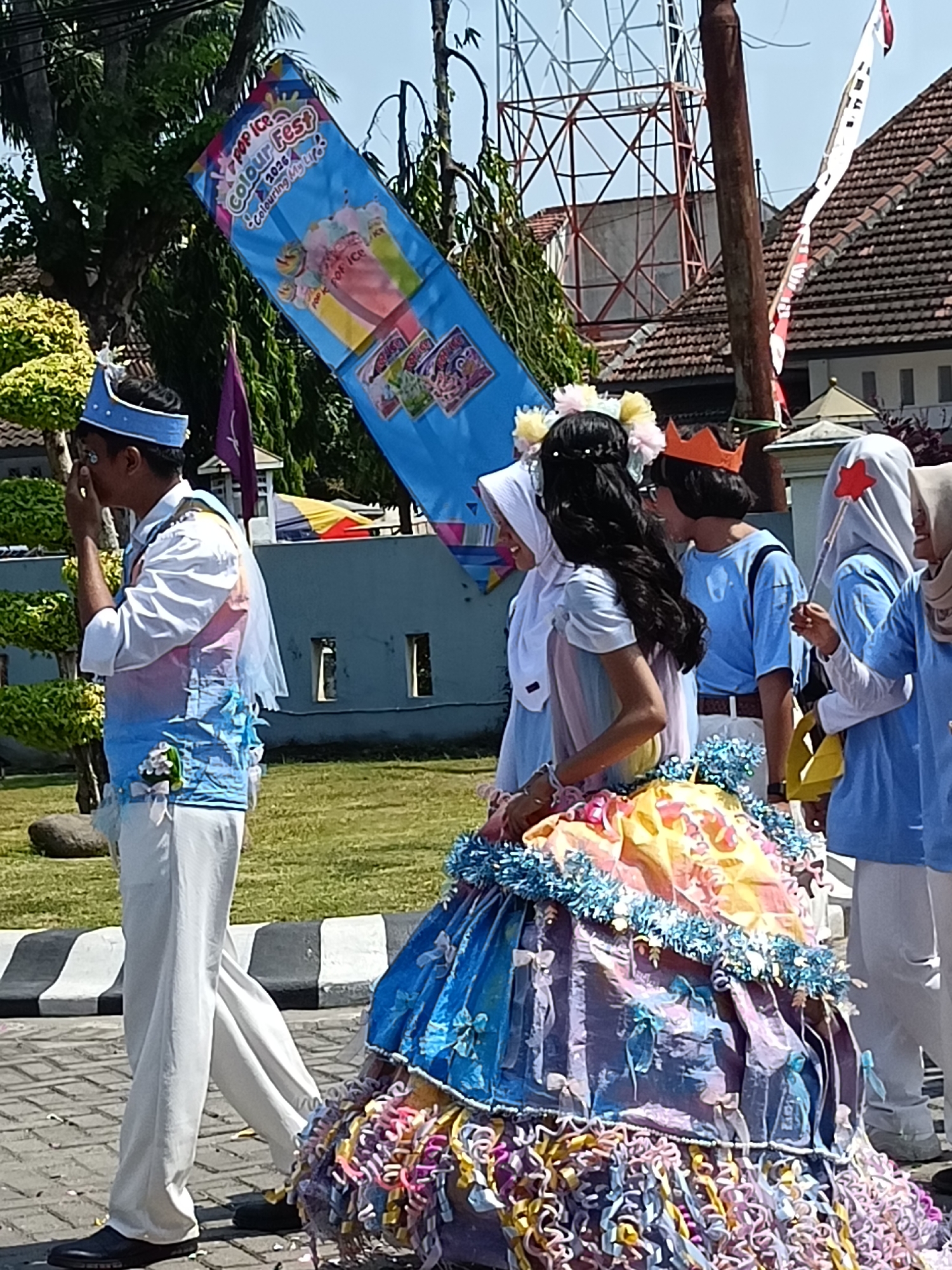 The image shows a group of people in what appears to be a parade or festival, possibly in Kediri City, Indonesia. A prominent blue banner with colorful images of ice cream and the text "POP ICE Colour Fest 2020 - Colouring My Life!" is displayed diagonally. In the foreground, a person in a white shirt, a light blue vest adorned with colorful decorations and a veil, and white pants, wearing a blue crown, is walking and touching their face. To their right, a person with dark hair, wearing a colorful, voluminous dress and a flower crown, is facing away from the camera. Behind them, several other individuals are visible, some wearing blue shirts and headscarves. The background includes trees, a building with a tiled roof, and a metal structure that could be a tower. The ground is paved with interlocking bricks.