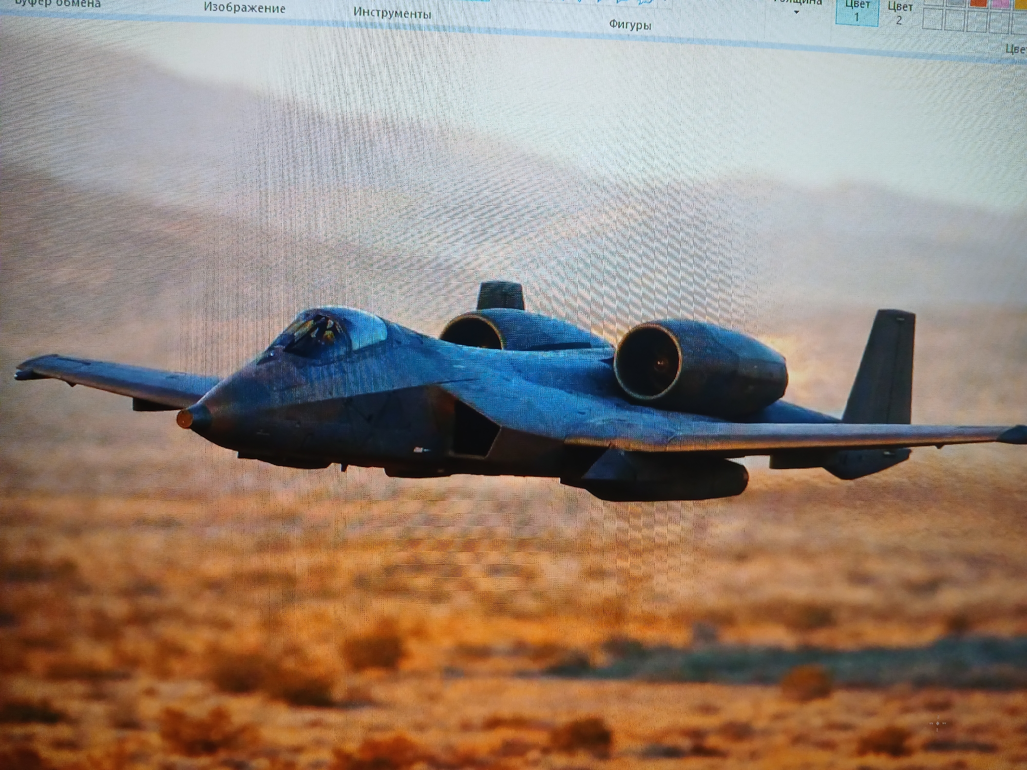 A dark-colored military aircraft is flying low over a desert-like landscape. The aircraft has two engines mounted high on the fuselage, a single vertical tail, and a large cockpit. The background is blurred, suggesting motion. The image appears to be captured from a computer screen, with program options visible at the top. The aircraft is not located in Kamianske, Ukraine, and is not flying over any urban environment.