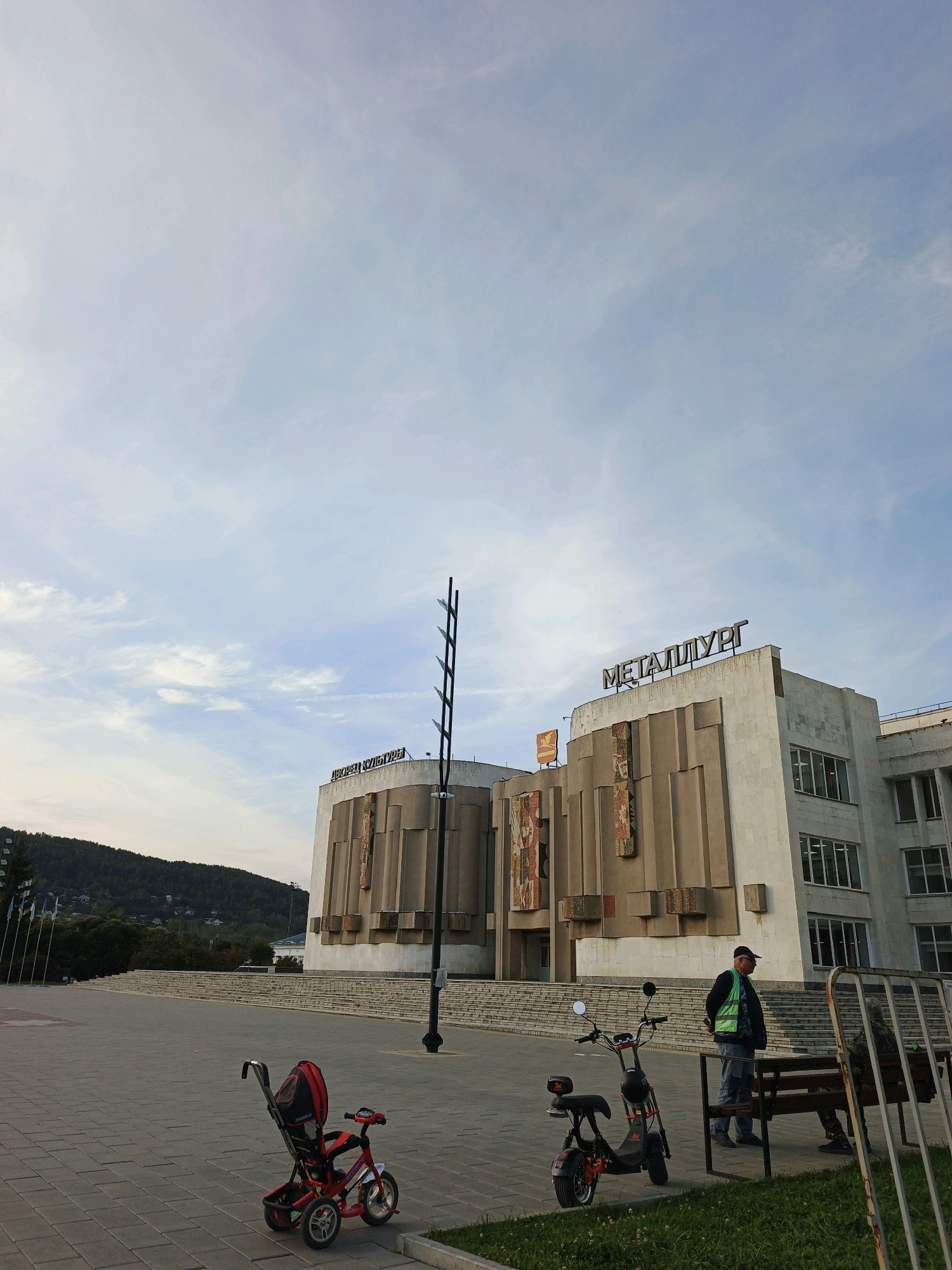 The image depicts the Palace of Culture "Metallurg" in Zlatoust, Russia, situated in a public square. In the foreground, a red child's tricycle is parked on a paved surface. To the right of the tricycle, a black electric scooter is also parked. A man wearing a green reflective vest and a baseball cap stands near the scooter. Further to the right, another person is seated on a park bench.
The "Metallurg" Palace of Culture is a prominent building with a distinctive facade featuring decorative mosaic panels and geometric concrete elements. A sign at the top of the building reads "МЕТАЛЛУРГ" (Metallurg). Adjacent to it, another sign reads "ДВОРЕЦ КУЛЬТУРЫ" (Palace of Culture). In the background, a hill covered in trees and dotted with houses rises, and several flags are visible on flagpoles to the left. The sky is partly cloudy. The scene suggests an outdoor public space with a focus on the cultural landmark.