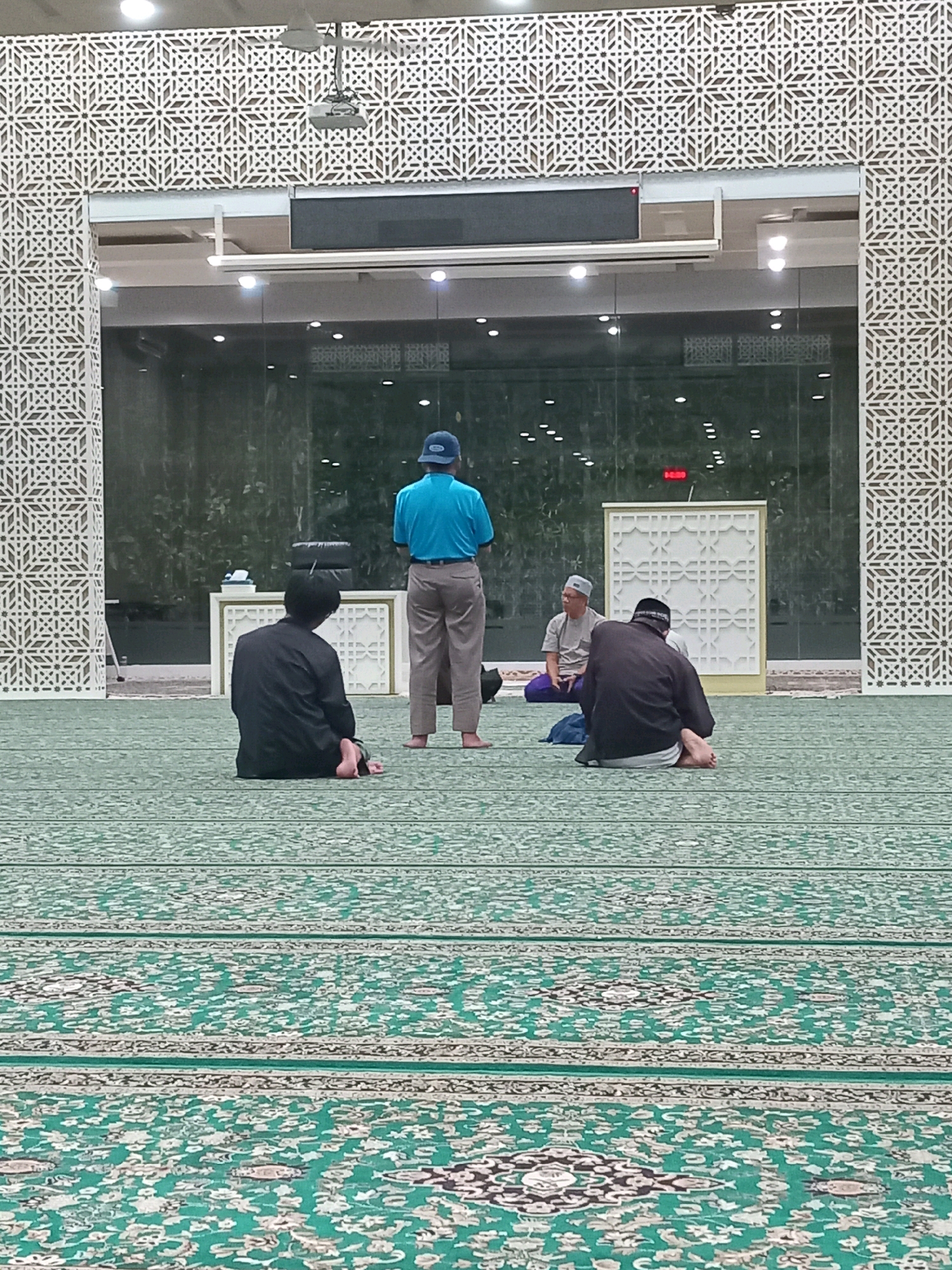 In Randuagung, Indonesia, inside a mosque, four individuals are present on a green carpet with a patterned design. Two men are seated facing away from the camera, with their backs to the viewer, on the carpet. One of these men is wearing a black garment, and the other is wearing a grey shirt and purple trousers. Another man is standing in the center of the frame, facing away from the camera, wearing a blue shirt and tan trousers, with a blue hat. A fourth man, also facing away from the camera, is partially visible behind a white podium with a decorative patterned screen. The mosque features geometric patterned walls and a large glass window or wall. A projector is mounted on the ceiling. The lighting appears to be artificial.