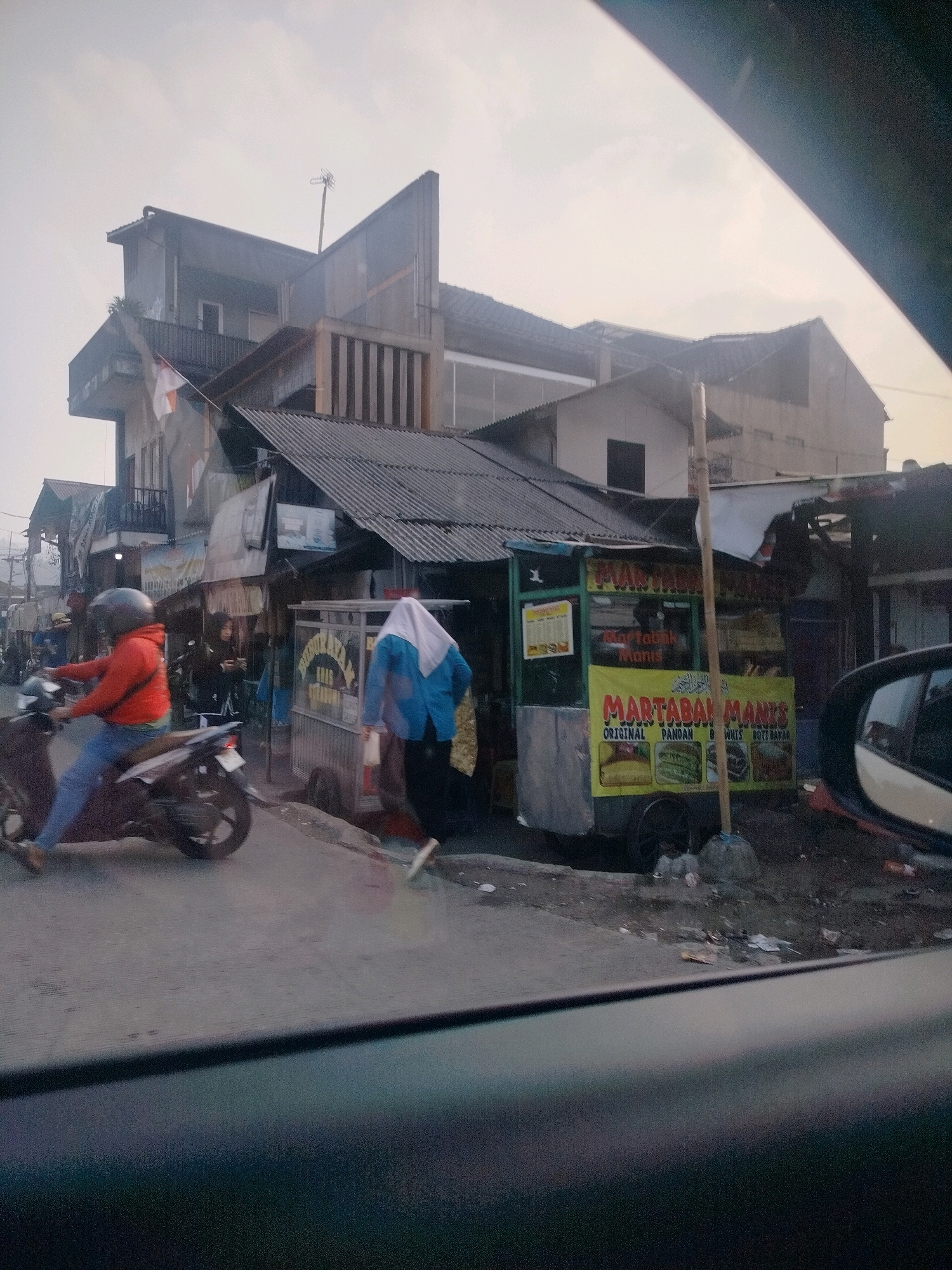 The image depicts a street scene in Megamendung, Indonesia, characterized by modest, multi-story buildings and roadside vendors. The sky is overcast. In the foreground, a motorcycle with a rider wearing a red jacket and a helmet is moving from left to right. To the right of the motorcycle, a person in a blue top and headscarf walks away from the camera. Behind them is a food cart labeled "MARTABAK MANIS" (sweet pancake), with various types of martabak advertised. Several other small stalls and buildings line the street, some with corrugated metal roofs. An Indonesian flag is visible on one of the buildings. The ground is paved, with some debris scattered along the roadside. A car's side mirror is partially visible on the far right of the frame.