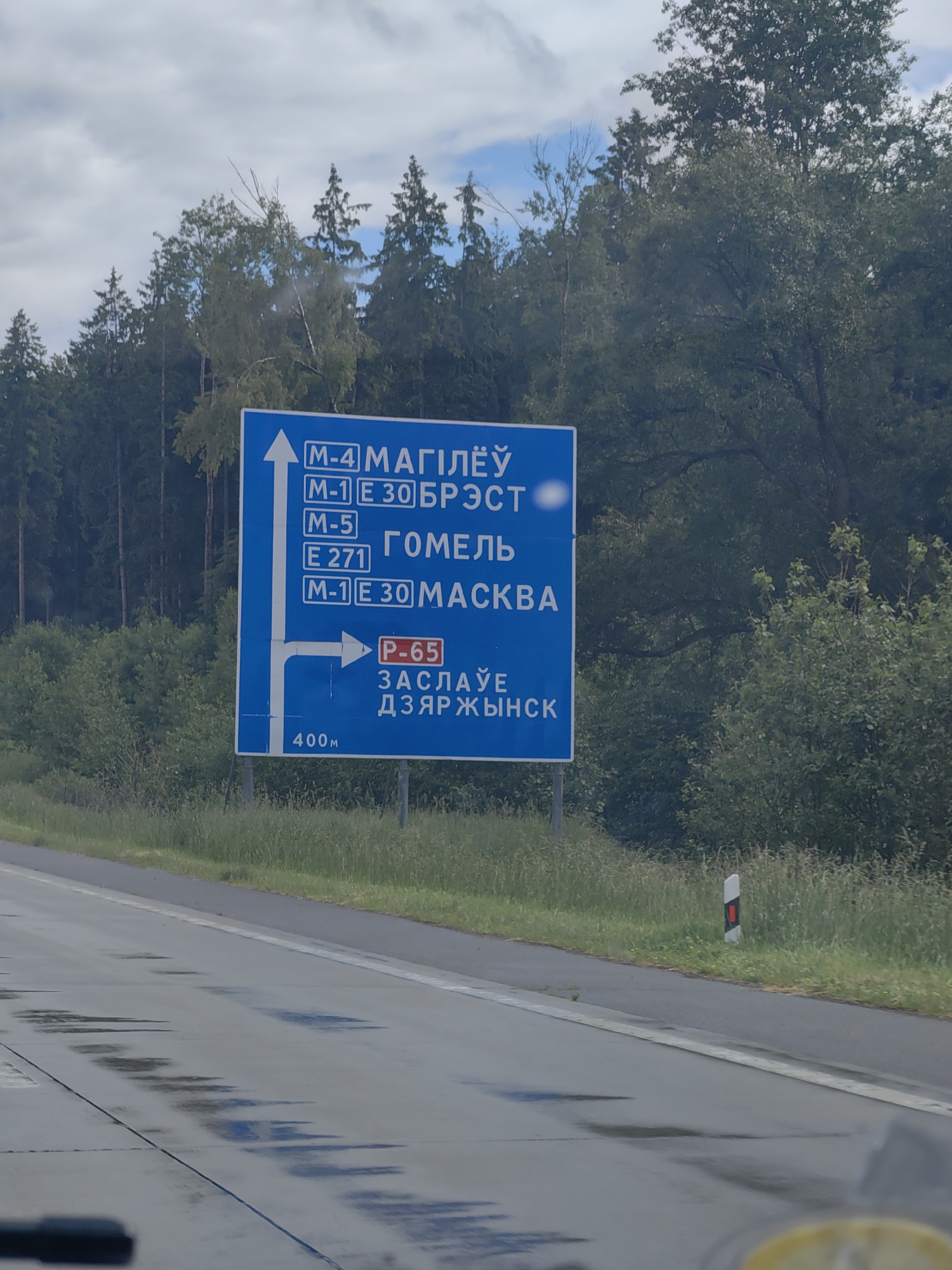 The image shows a blue road sign in the foreground with white text and arrows. The sign lists destinations and route numbers. The top section indicates an upward arrow with "M-4 МАГІЛЁЎ" and "M-1 E 30 БРЭСТ". Below that is "M-5" followed by "ГОМЕЛЬ". The next line reads "E 271" and "M-1 E 30 МАСКВА". The lower part of the sign shows a right-pointing arrow and "P-65" in a red rectangle, followed by "ЗАСЛАЎЕ" and "ДЗЯРЖЫНСК". At the bottom of the sign, "400м" is written. To the left of the main sign is a smaller blue sign with a white arrow pointing upwards and left. The road surface in the foreground is wet, with visible puddles and reflections. A white line marks the edge of the road. Beyond the road, there is a grassy verge with a few small yellow flowers, and then a dense line of trees, primarily coniferous, with some deciduous trees interspersed. The sky is overcast with patches of blue sky visible. In the distance, to the right of the road, a white and red road marker is visible. The image appears to be taken from a moving vehicle, looking towards the road sign and the surrounding environment. This road sign is located on the M4 highway, likely indicating directions to Mogilev, Brest, Gomel, Moscow, Zaslavl, and Dzerzhinsk. Given the presence of "ЗАСЛАЎЕ" and "ДЗЯРЖЫНСК", the location is likely near these areas within the Путчынскі сельскі Савет in Belarus, as these are prominent destinations indicated by the sign.
