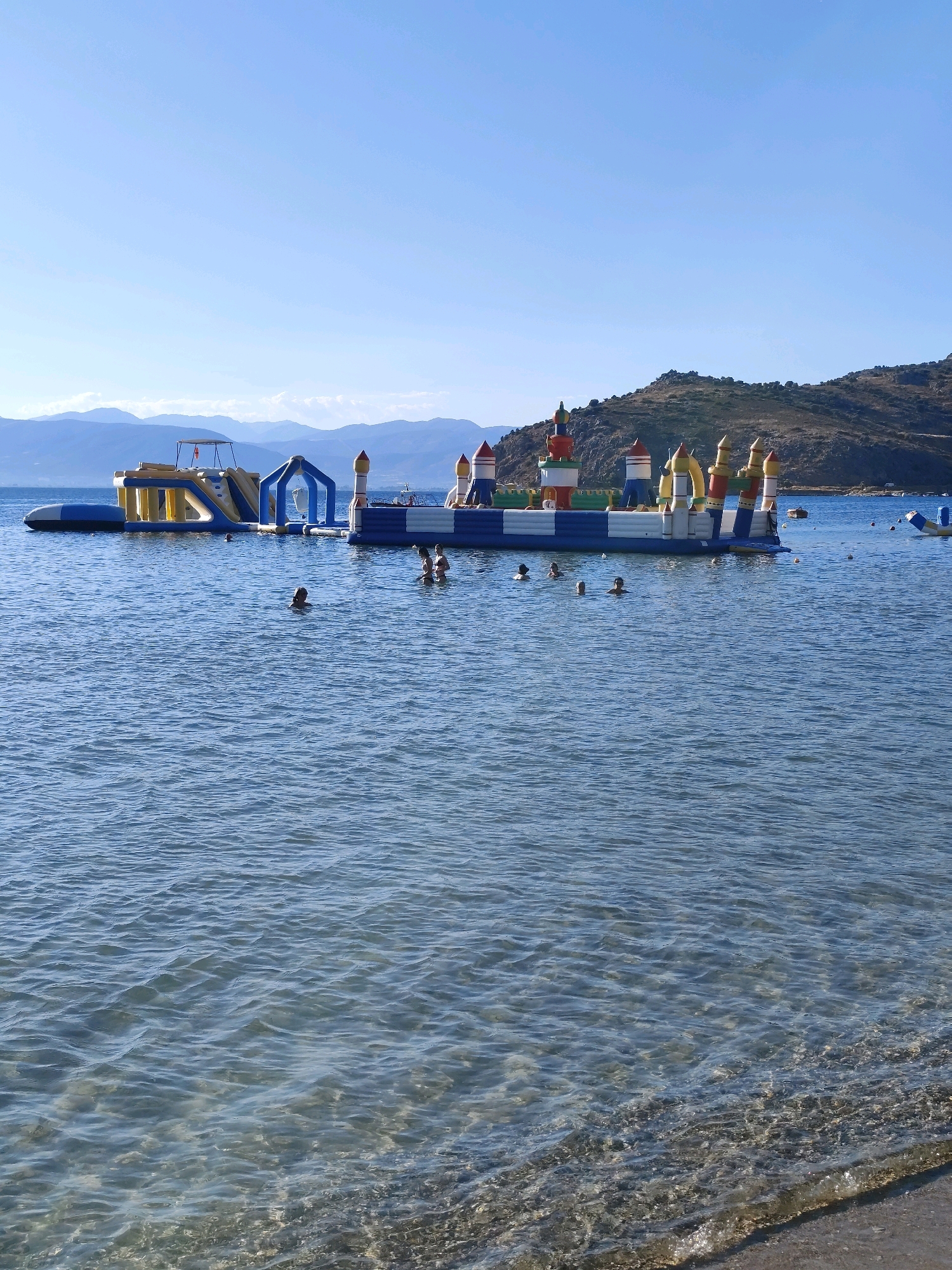 The image shows a vibrant inflatable water park with slides and towers floating on the calm, clear blue waters of the sea. The park is situated in the Municipal Unit of Nafplio, Greece, with a picturesque backdrop of rolling green hills and mountains under a clear blue sky. Several people are seen swimming and enjoying the water around the inflatable structures. The foreground features the shallow, pebbled shore, with gentle waves lapping at the edge. The sunlight reflects off the water, creating a serene and inviting atmosphere for a summer day at the beach.