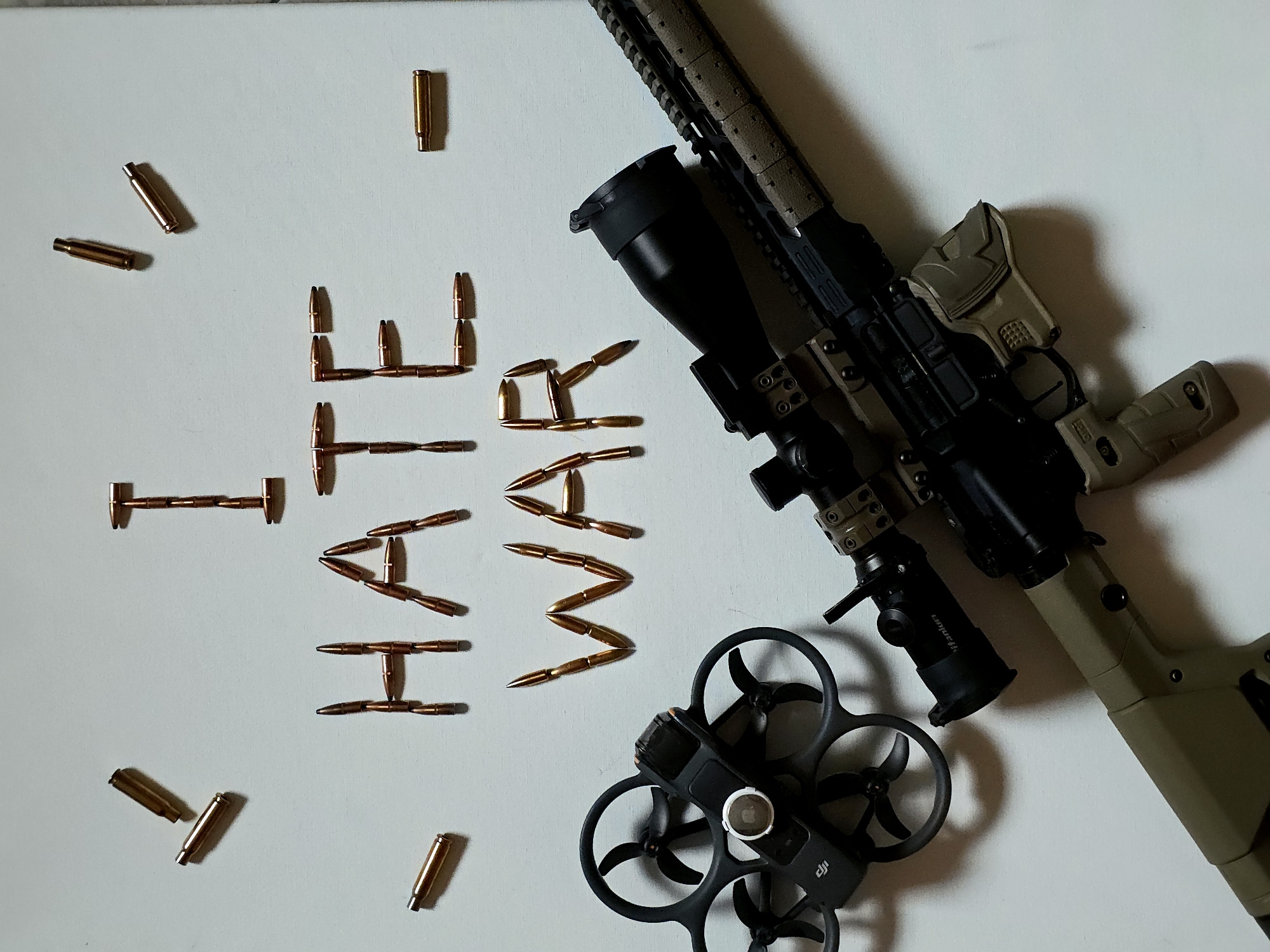 The image displays a stark still-life composition on a plain, light-colored surface. The central focus is the phrase "HATE WAR," meticulously spelled out using numerous brass rifle cartridges. The word "HATE" is positioned on the left, formed with cartridges arranged horizontally and vertically, while "WAR" is on the right, also constructed from individual cartridges. Scattered around these words are additional loose cartridges.
To the right side of the frame, a modern rifle is partially visible, featuring a tan and black finish. It is equipped with a prominent black scope, which bears the faint text "Phantom" on its side. Below the rifle, a black DJI Avata drone rests on the surface, identifiable by its distinctive ducted propellers and central camera housing. The "DJI" logo and "Avata" text are visible on its body. The scene is illuminated by soft, even lighting, suggesting an indoor setting. The arrangement of weaponry and a modern drone alongside an explicit anti-war message creates a powerful and thought-provoking statement.