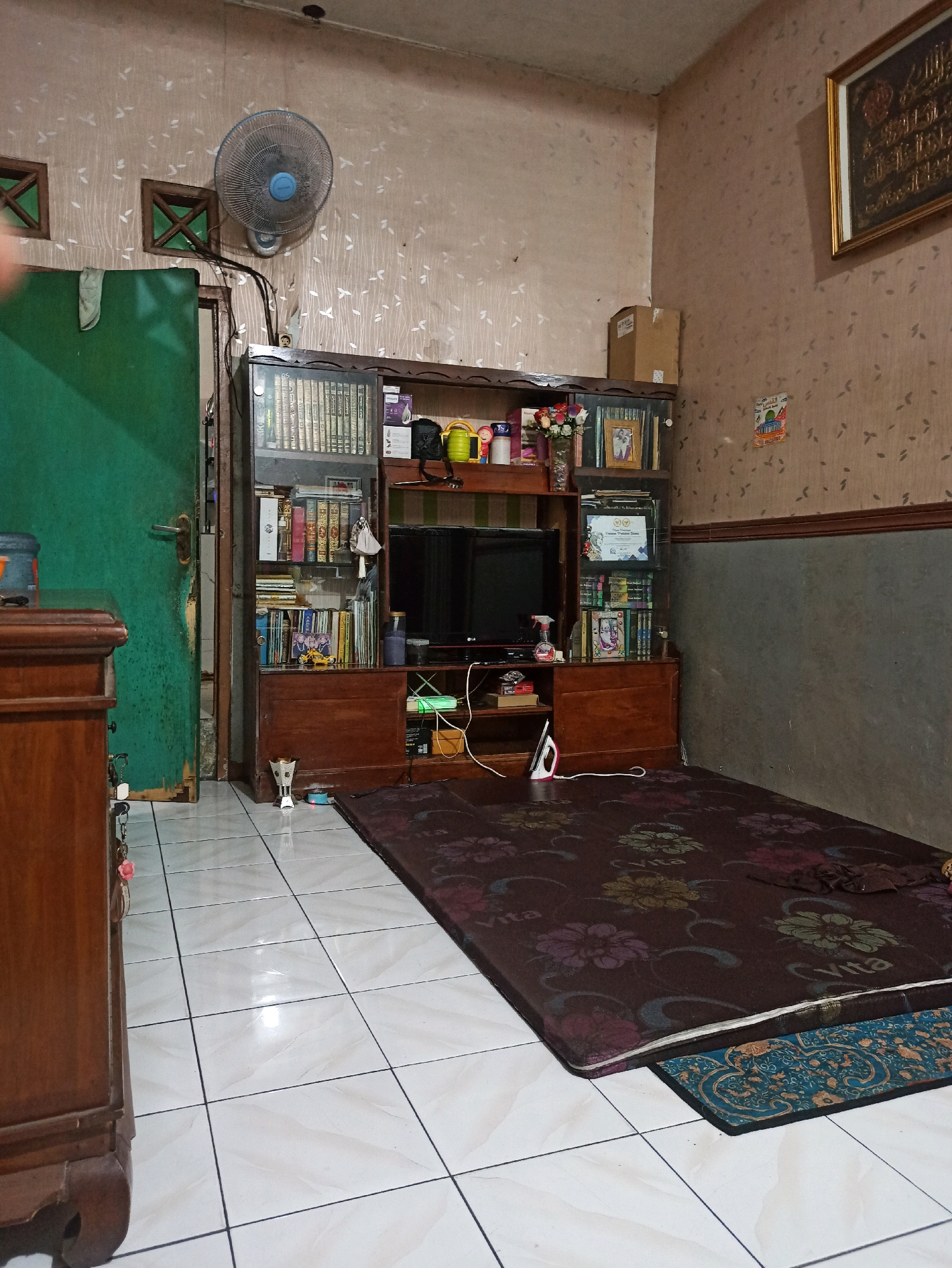 The image depicts the interior of a room, possibly a living room or bedroom, in North Jakarta, Indonesia. The scene is cluttered with various objects, suggesting a lived-in space. Dominating the center of the frame is a wooden entertainment center filled with books, decorative items, and a television. To the left, a tall green wooden door is partially visible, with a fan mounted on the wall above it. The floor is tiled with light-colored square tiles. A large, dark-colored mattress with floral patterns lies on the floor in the foreground, partially covering a smaller patterned rug. The walls are covered with textured wallpaper, and a framed piece of artwork hangs on the right wall. A wooden cabinet is partially visible on the far left. The lighting appears to be artificial and somewhat dim, casting shadows in some areas.