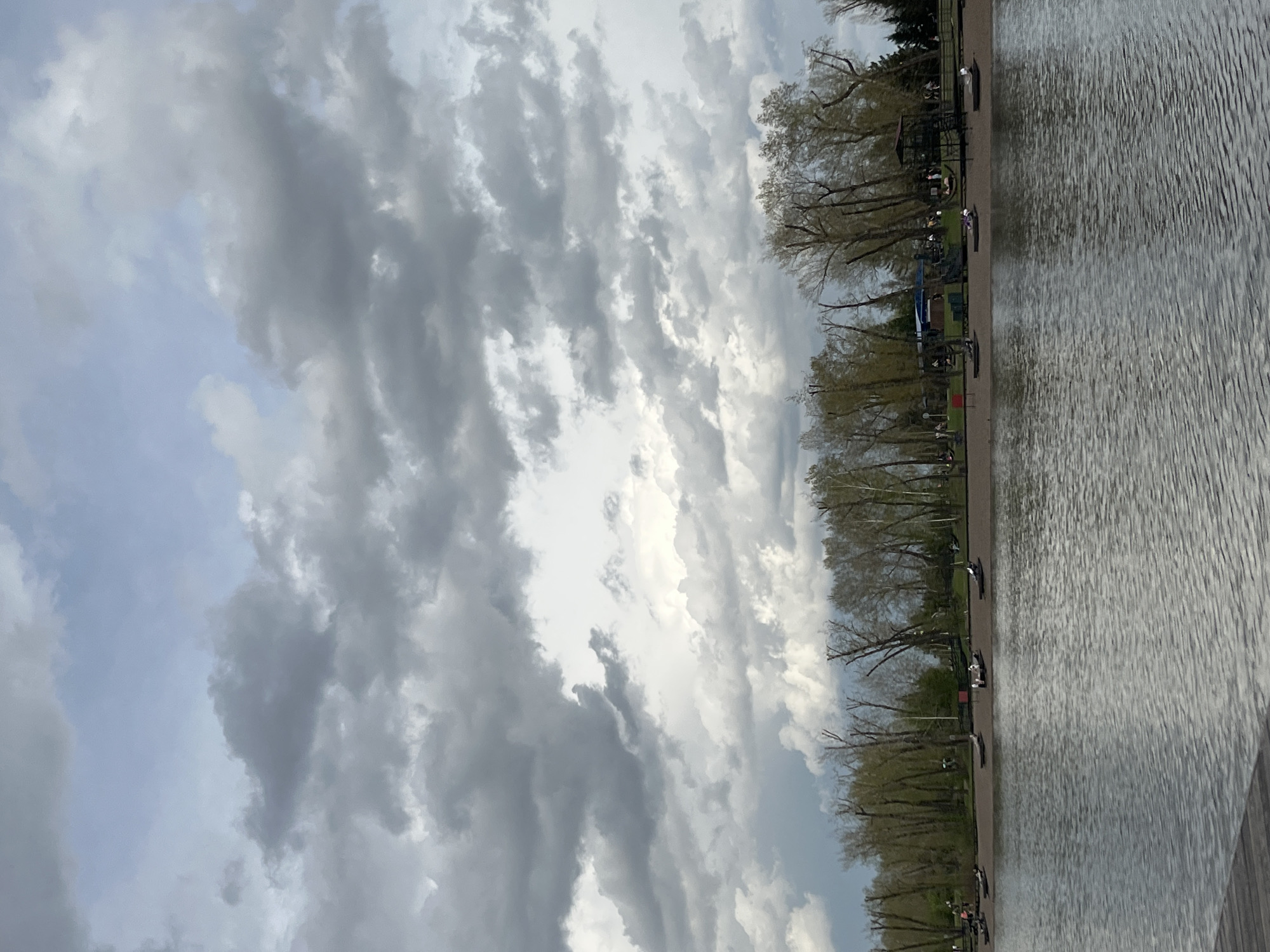 The image depicts a waterfront scene, possibly on the Yenisey River, in Krasnoyarsk, Russia. The sky is filled with dramatic, layered clouds. The river's surface is rippled, indicating wind. The shoreline is lined with trees and what appears to be a paved walkway, where individuals can be seen at various points. Small boats or kayaks appear to be present along the shore. The overall environment conveys an outdoor recreational area along a riverfront setting.