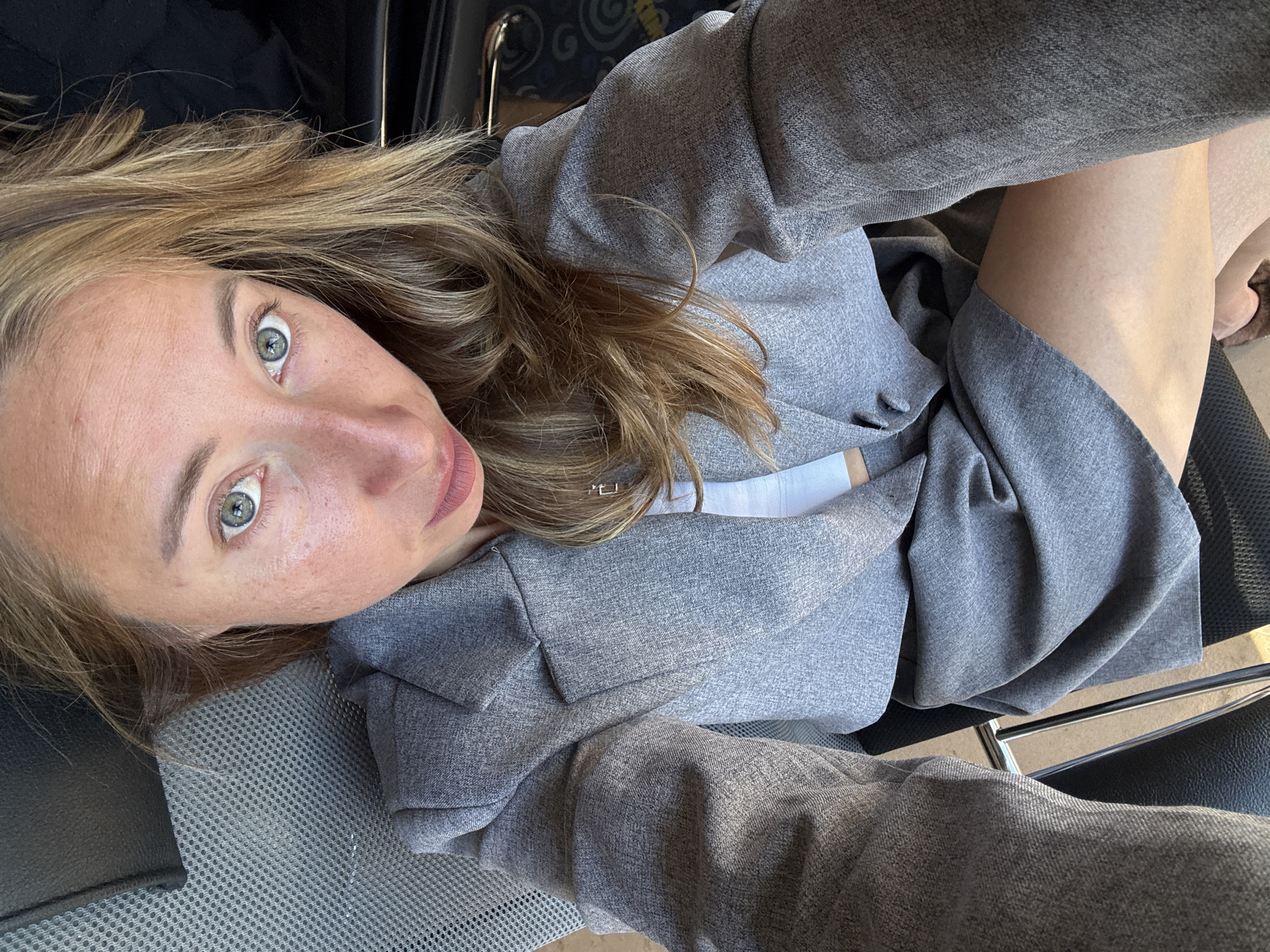 A woman with blonde hair and light green eyes is captured in a selfie, looking directly at the camera. She is wearing a grey blazer over a white top, and her legs are visible. The background appears to be an indoor setting, possibly an office or a public transport seat, given the presence of textured grey fabric and dark upholstery. The lighting suggests it's daytime, with sunlight casting shadows and highlighting her features. No specific landmarks in Chaikovsky, Russia are identifiable from the image.