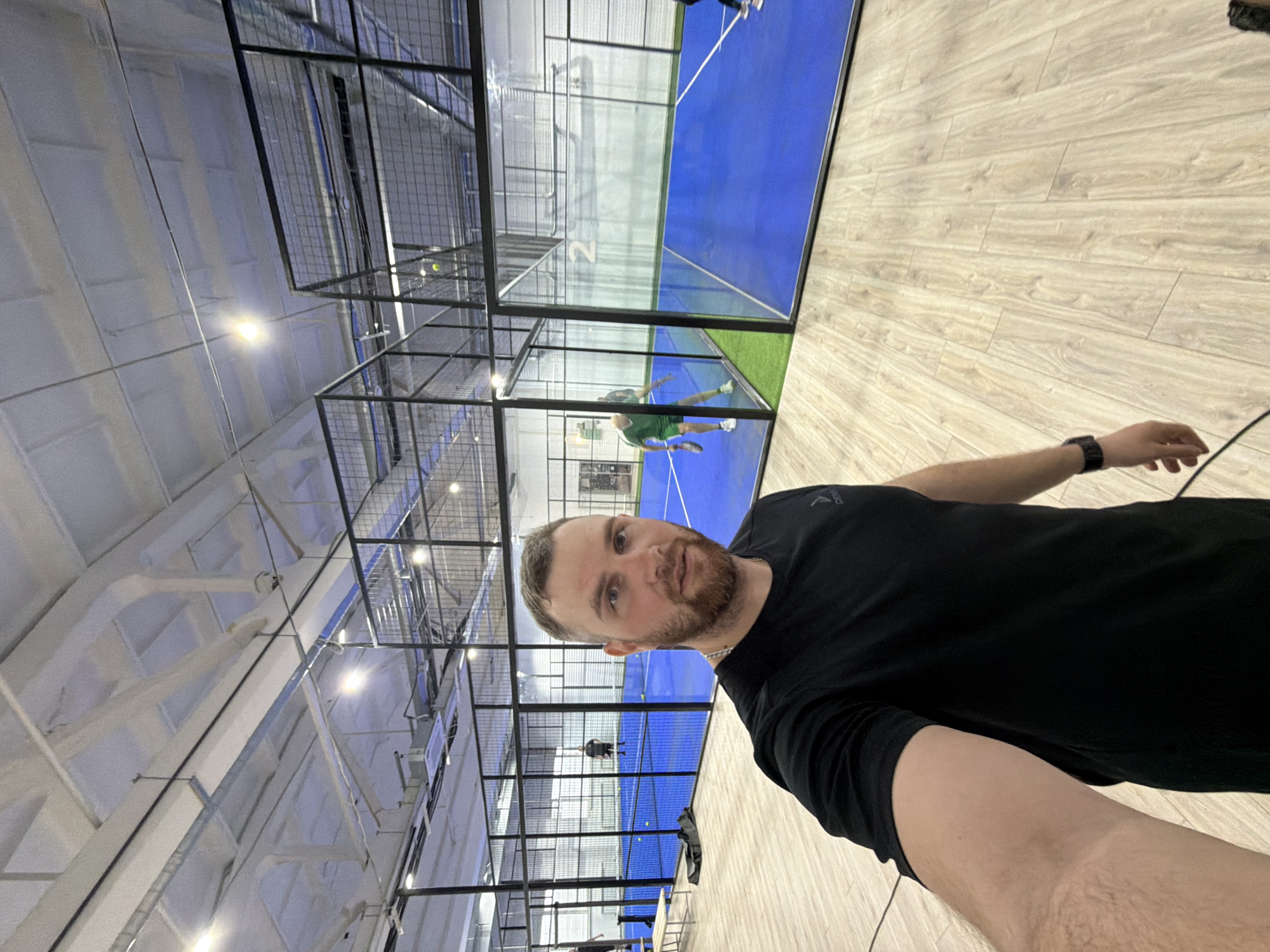 The image shows a man taking a selfie indoors at a padel tennis facility. He has light skin, a beard, and is wearing a black t-shirt and a smartwatch. In the background are multiple padel tennis courts with blue playing surfaces enclosed by black metal mesh. Other people are visible playing on some of the courts. The facility has a high ceiling with exposed metal beams and lighting fixtures. The floor is made of light-colored wood. I am unable to locate the city of Chaikovsky, Russia, due to it not being indexed by Google Maps.