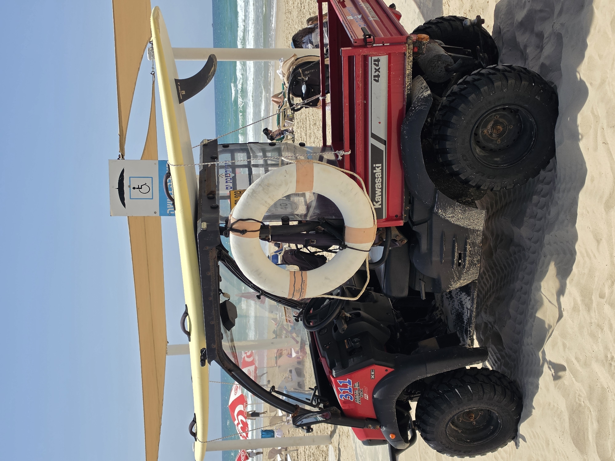 A red Kawasaki Mule SX 4x4 utility vehicle is parked on a sunny beach, likely in Bat Yam, Israel, serving as the central subject of the image. The vehicle is equipped with a yellow surfboard strapped to its side and a white lifebuoy, marked with "40", attached to the front roll bar.
In the background, the clear blue Mediterranean Sea stretches out, with gentle waves breaking on the sandy shore. Several people are visible relaxing on the beach under colorful umbrellas and swimming in the ocean, creating a leisurely atmosphere. A light-colored canopy extends over the utility vehicle, providing shade.
A white sign with a blue border is prominently displayed, featuring a wheelchair accessibility symbol and another symbol depicting what appears to be a person or a bird under an umbrella shape. Hebrew text on the sign includes "מירבית" (Meravit / maximum) and "שבת ים" (Shabbat Yam / sea weekend). Additional, partially visible Hebrew text is present.
The scene depicts bright daylight under a clear blue sky, suggesting good weather conditions. Visible text on the Kawasaki vehicle includes "311", "MULE SX", "XC", "Kawasaki", and "4x4". A tire shows "URD" (likely part of a brand name). Other indistinct numbers and letters are visible on stickers on the vehicle.