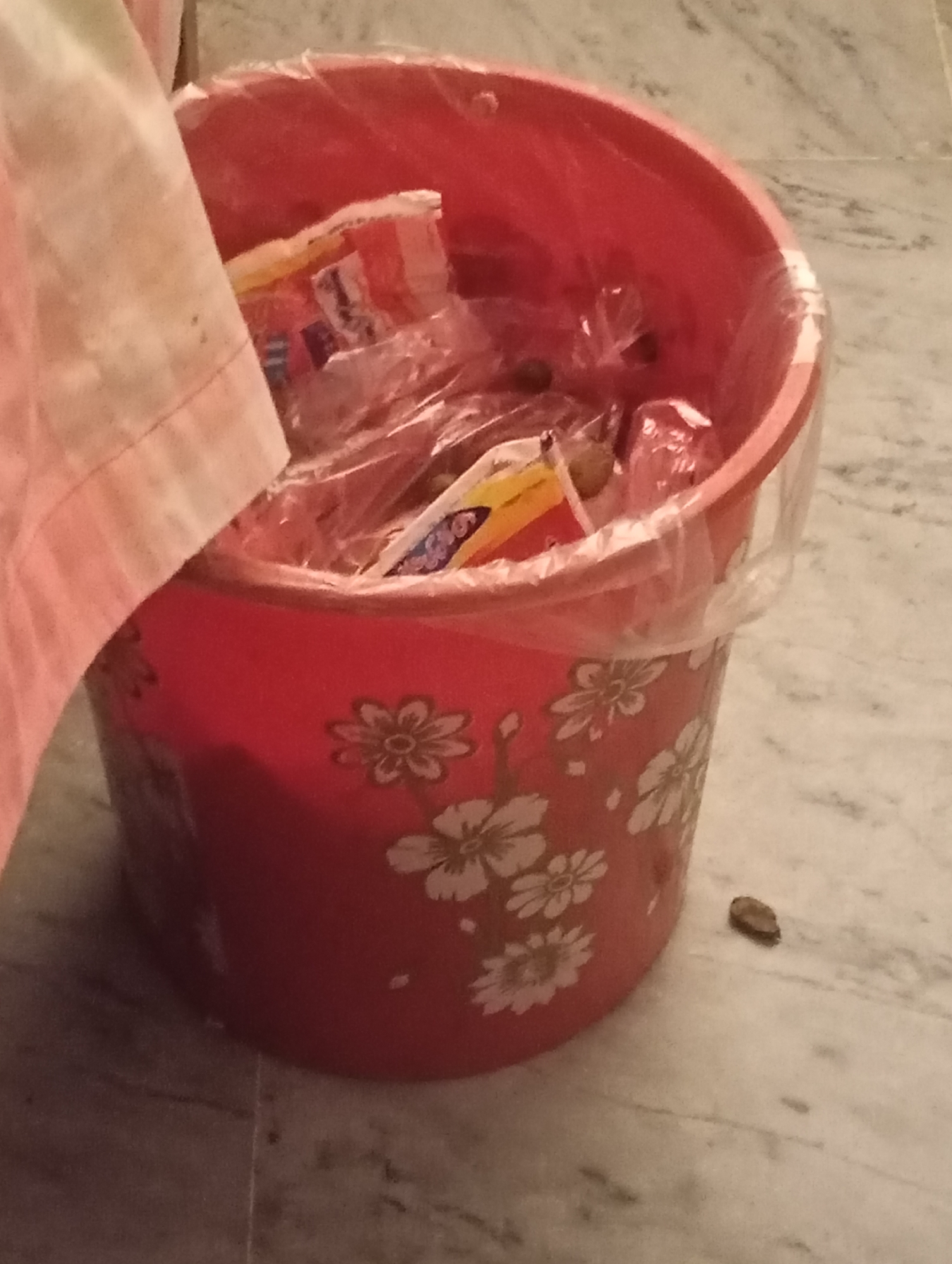 A medium shot shows a red plastic trash can with white floral patterns on the side. The trash can is filled with various items wrapped in clear plastic, some of which appear to be snack wrappers. A pink and white striped cloth is draped over the left side of the trash can, partially obscuring its contents. The trash can is situated on a light-colored marble floor, and a single small, brown object, possibly a seed, is visible on the floor to the right of the trash can. The lighting is dim, creating shadows on the floor.