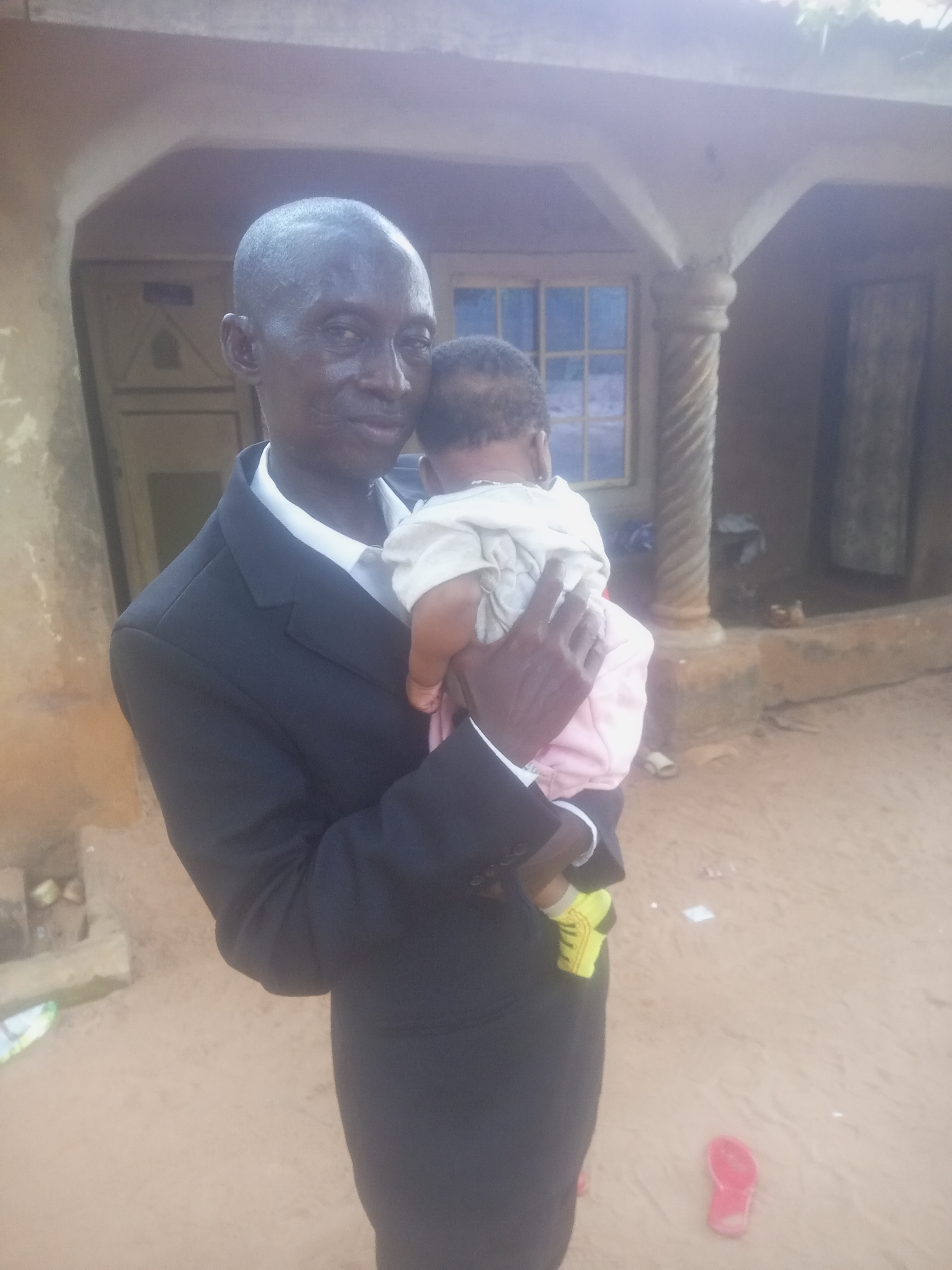 A man in a black suit and white shirt holds a baby wrapped in a light-colored cloth. The man is bald and has dark skin. The baby is also dark-skinned and appears to be wearing a pink garment. They are standing in front of a building with a rough, textured exterior. The building has a covered porch with a decorative column. Behind the porch, a window with multiple panes is visible. The ground is covered in sand or dirt. A red sandal lies on the ground in the foreground.