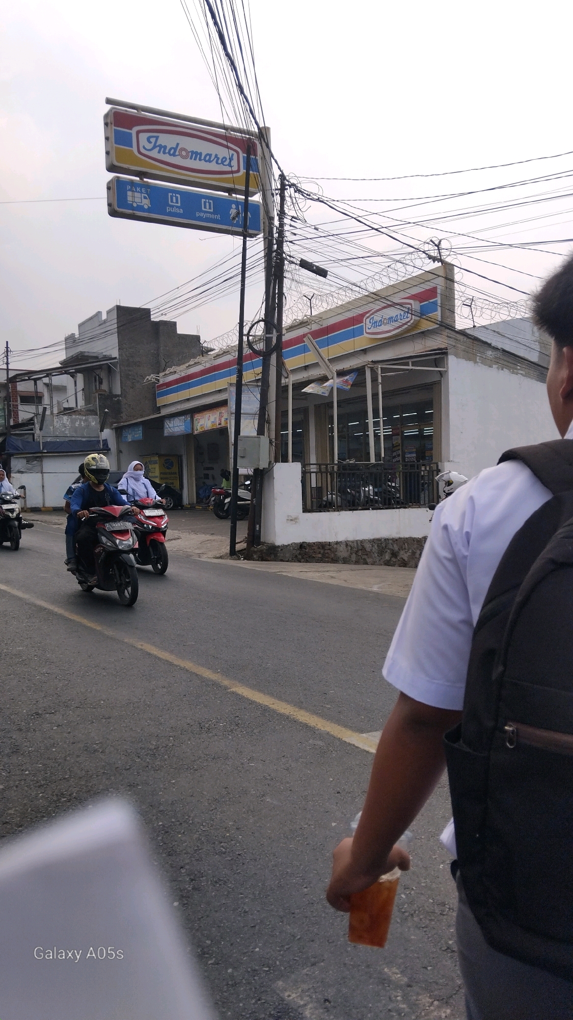 The image shows a street scene in Sumedang, Indonesia, with a prominent Indomaret convenience store on the right. The store has a white facade with red, blue, and yellow stripes across the top. A large sign for Indomaret is visible both on top of the store and on a pole on the left side of the image, which also displays icons for "paket," "pulsa," and "payment."
Several motorcycles are on the road. Two people are riding on a red motorcycle in the foreground, with the rider wearing a yellow helmet and the passenger wearing a white headscarf. Another motorcycle with two people is visible behind them, also with a passenger in a white headscarf. A person is walking on the sidewalk on the right, from the rear view, wearing a white shirt and a backpack. They are holding a clear plastic cup with a dark-colored beverage.
The street is asphalt with a yellow dividing line. Buildings line the left side of the street, appearing to be part of a commercial or residential area. Power lines are strung across the sky above the street and buildings. The sky is overcast. The overall environment appears to be a typical urban street in Indonesia. The bottom left corner of the image shows "Galaxy A05s" indicating the device used to capture the photo.