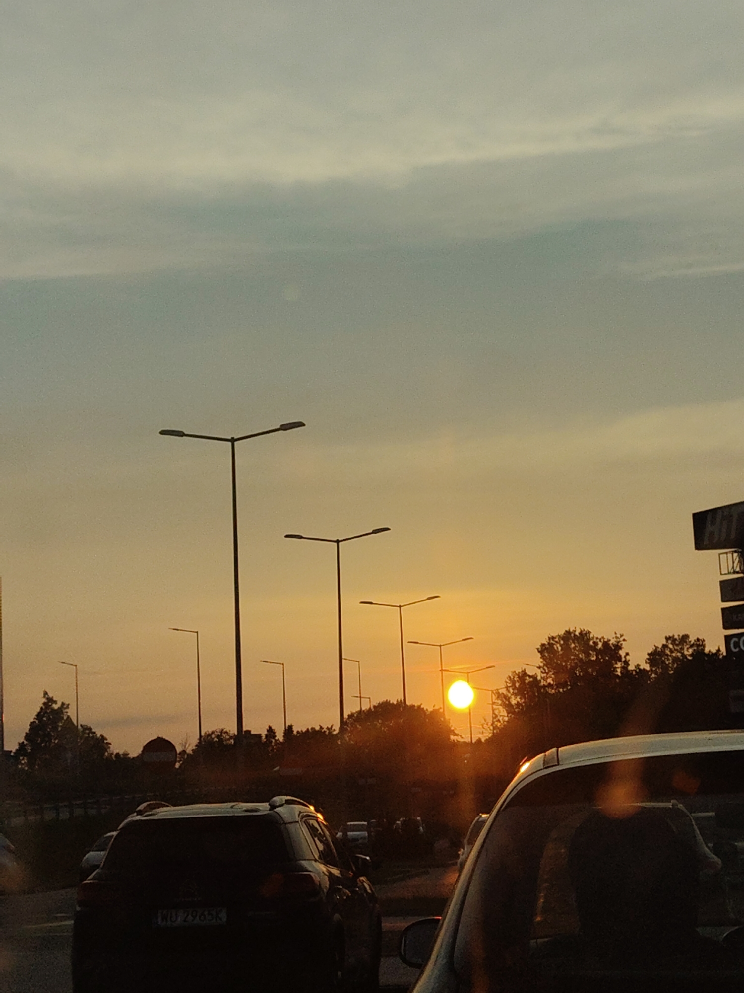 The image shows a sunset over a road in Nowy Dwór Mazowiecki, Poland. The sky is filled with warm orange and yellow hues, with some lighter clouds visible. Several tall lampposts with multiple light fixtures are spaced along the road. In the foreground, a dark-colored SUV is visible, with its license plate showing "WU 2965K". To the right, a tall sign with the word "HIT" is partially visible, along with other signage. Trees line the side of the road, silhouetted against the bright sunset. The sun is a bright, glowing orb near the horizon, casting a warm light and lens flares across the scene. Traffic appears to be moving slowly or stopped, as suggested by the presence of multiple vehicles in the frame.