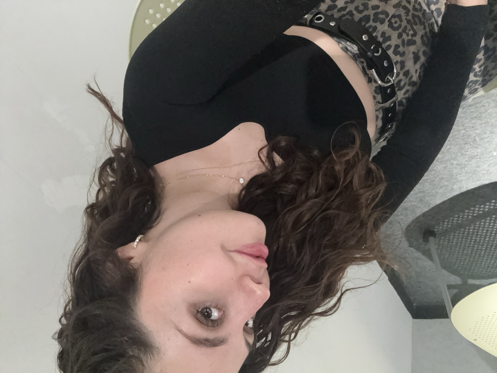 A woman with curly brown hair is centered in the image. She is wearing a black long-sleeved shirt with a cutout at the chest. A leopard print skirt or pants is visible, secured with a black belt with silver hardware. A gold necklace with a small pendant is around her neck. She is wearing a small earring on her left ear. She appears to be indoors, seated on a chair with a perforated pattern. In the background, there is a light-colored wall. There is no information in the image to indicate that this is Mexico City, Mexico.