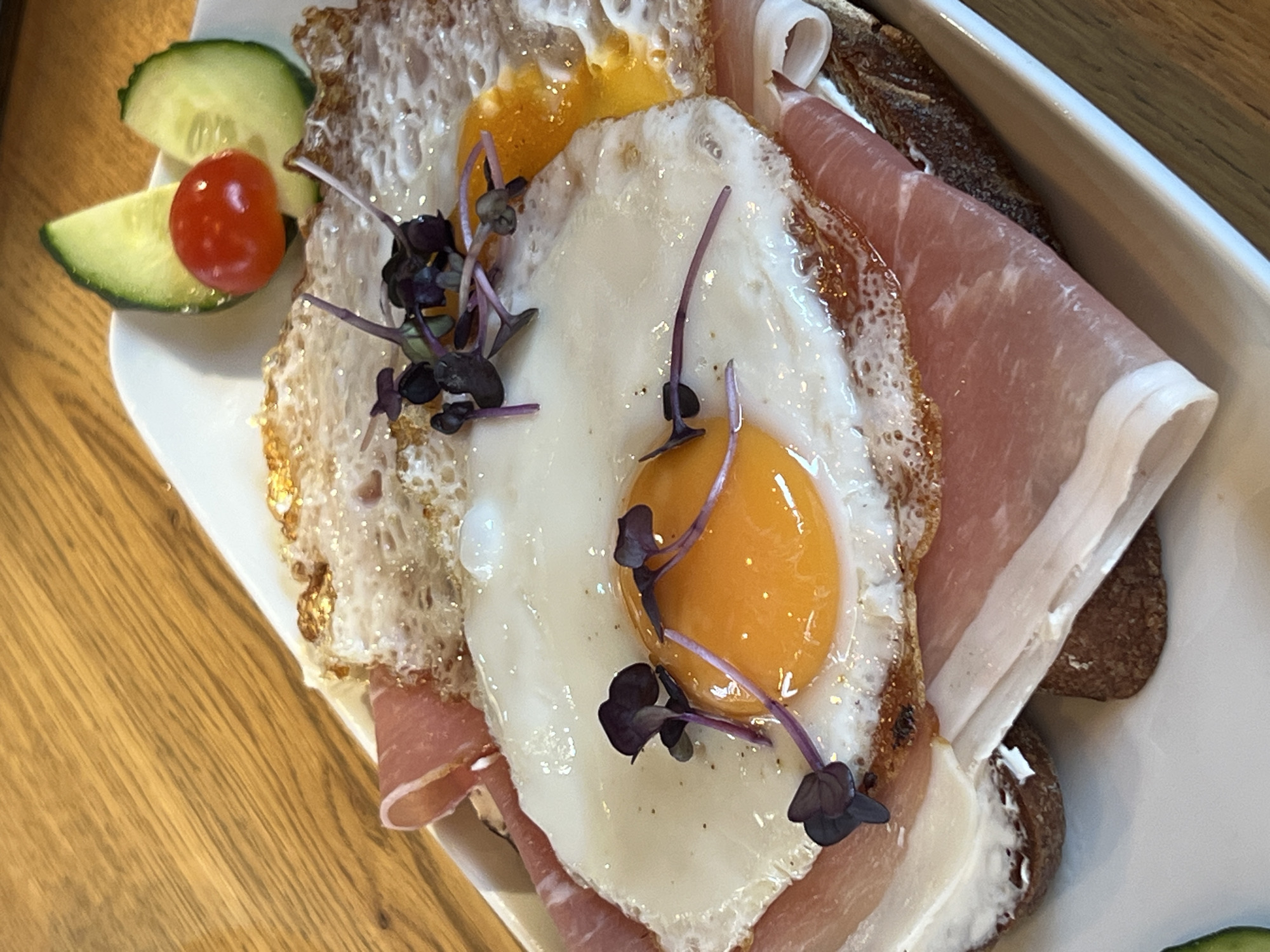 A close-up, overhead shot shows a white rectangular plate with a breakfast dish arranged on it, resting on a wooden surface. The dish features a slice of dark bread topped with what appears to be cream cheese, thin slices of cured ham, and two fried eggs. The eggs have bright yellow yolks and crispy white edges. Small, dark purple sprouts are scattered over the eggs. To the upper right of the dish, on the plate, are two thin slices of cucumber and one small red cherry tomato. The background shows a textured wooden tabletop. The overall impression is of a single serving of a savory breakfast. The location is a restaurant in Moosburg, Germany.