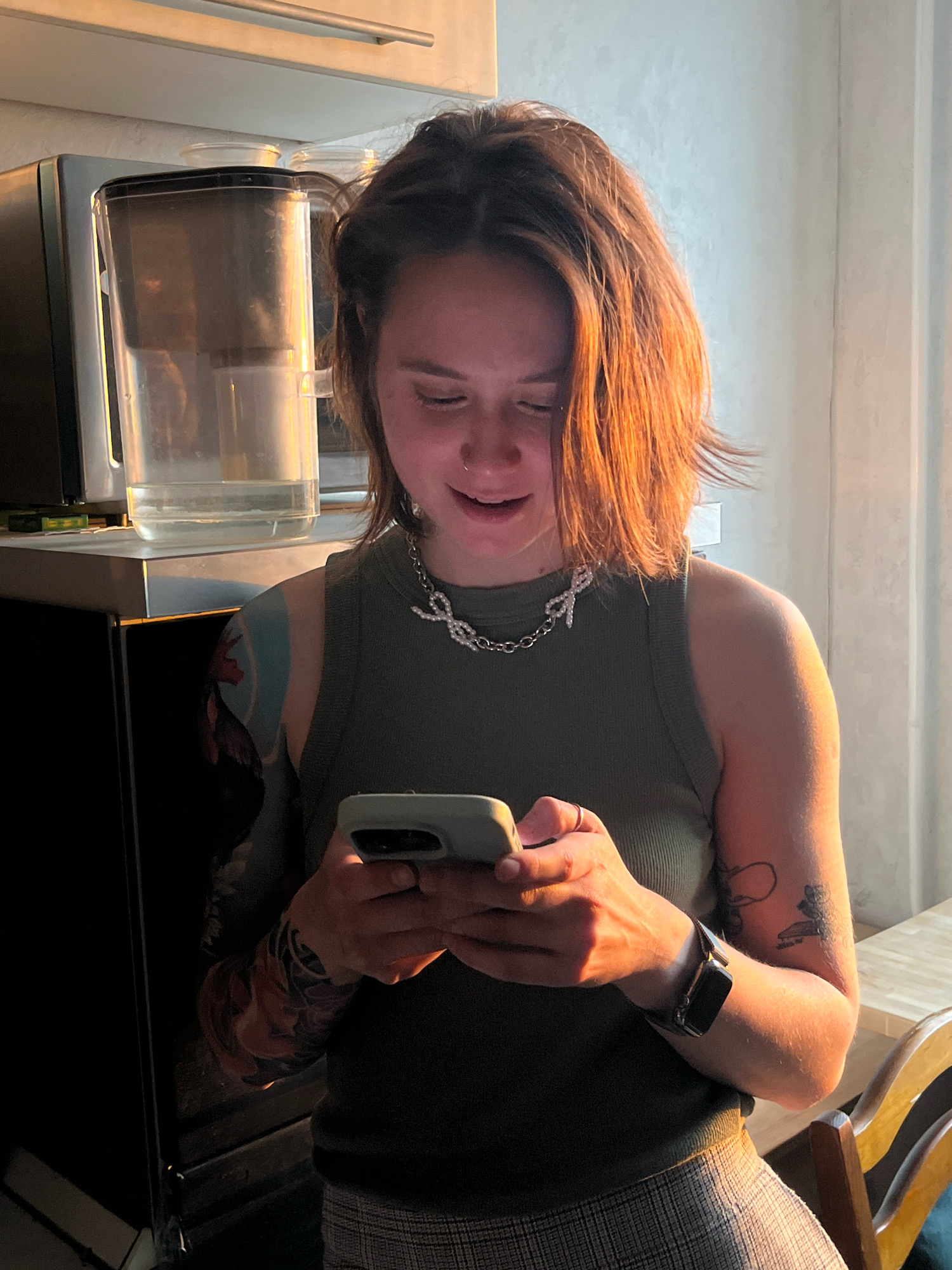 A young woman with auburn hair is looking down at her smartphone, illuminated by warm, low-angled sunlight. She is wearing a dark green, sleeveless, ribbed tank top and a silver necklace with a bow pendant. Her left arm is adorned with a large, colorful tattoo of a rooster. A ring is visible on her right index finger. She is standing in what appears to be a kitchen, with a water filter pitcher visible on a shelf behind her, next to a dark appliance. The background includes a light-colored wall and window with blinds.