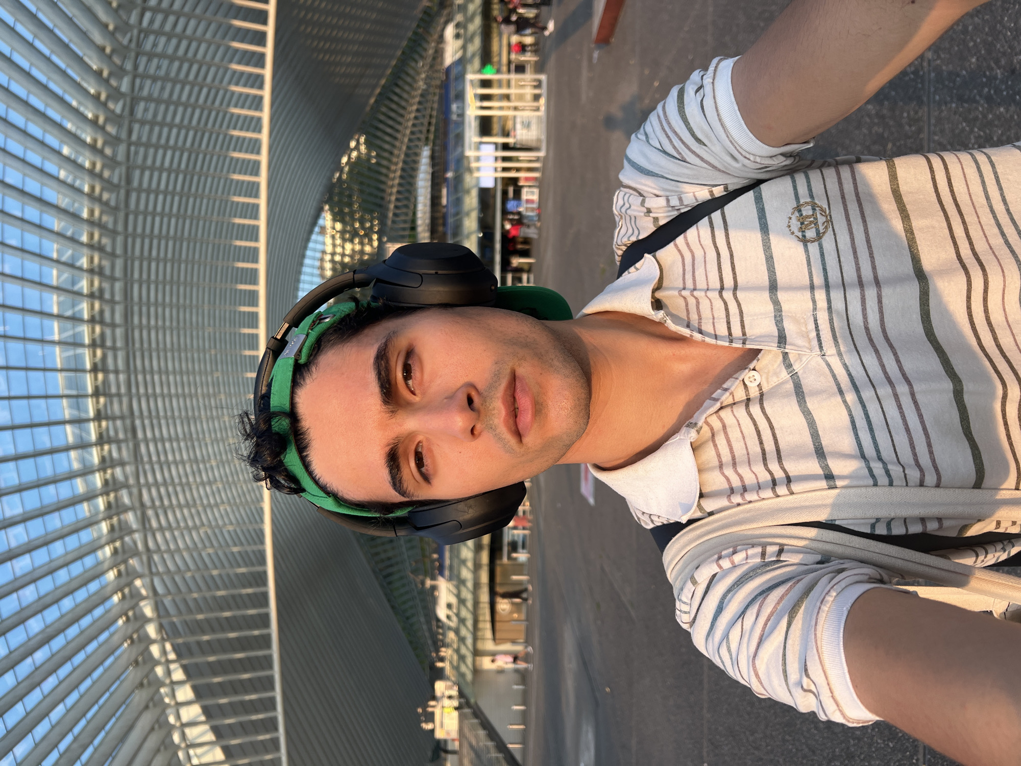 In the city of Liège, Belgium, a young man is taking a selfie in front of the Liège-Guillemins train station, an architectural marvel designed by Santiago Calatrava. The man, wearing a green cap, headphones, and a striped polo shirt, stands in the plaza outside the station. Sunlight casts long shadows across the ground, illuminating the modern, sweeping lines of the station's roof. People are visible in the background, suggesting a bustling transit hub. The man's expression is neutral as he captures the moment, likely enjoying the ambiance of the station and the warm, late-afternoon light.