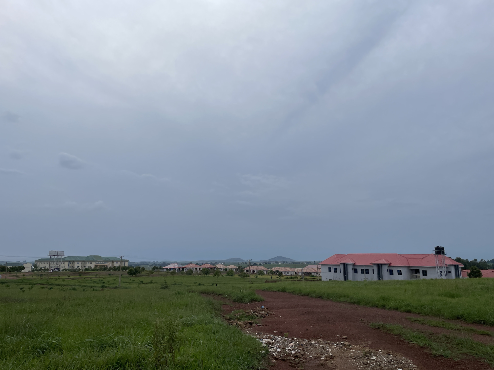 A partially constructed residential area is depicted under a cloudy sky in Durumi, Nigeria. In the foreground, a dirt path winds through a field of tall, green grass. To the right, a modern building with a red roof and white walls is visible, equipped with a large black water tank on a metal tower. In the middle ground, a row of similarly constructed houses with red roofs stretches across the landscape, interspersed with scattered trees. In the background, a larger, more institutional building with a green roof and a water tower stands prominently. The overall impression is one of ongoing development in a semi-rural setting.