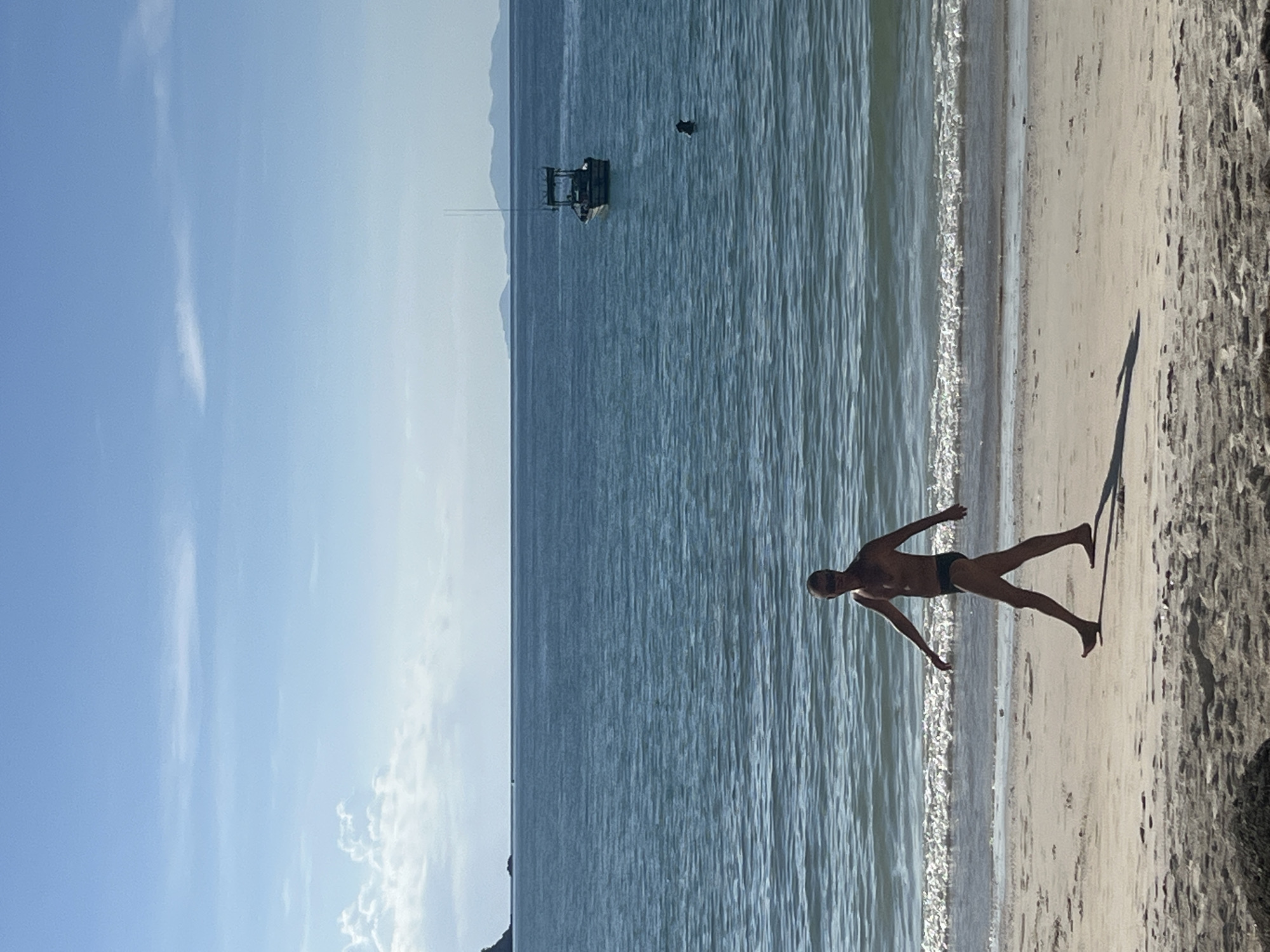 A man wearing sunglasses and black swim trunks walks along a sandy beach. He is facing to his left, with his right leg forward and his arms extended slightly. The ocean is calm and blue behind him, with small waves lapping at the shore. A boat is visible in the distance on the water, as are faint outlines of islands. The sky is clear and blue with a few scattered clouds. The sand is light brown with some darker patches and pebbles. This scene appears to be in ตำบลเกาะพะงัน, Thailand.