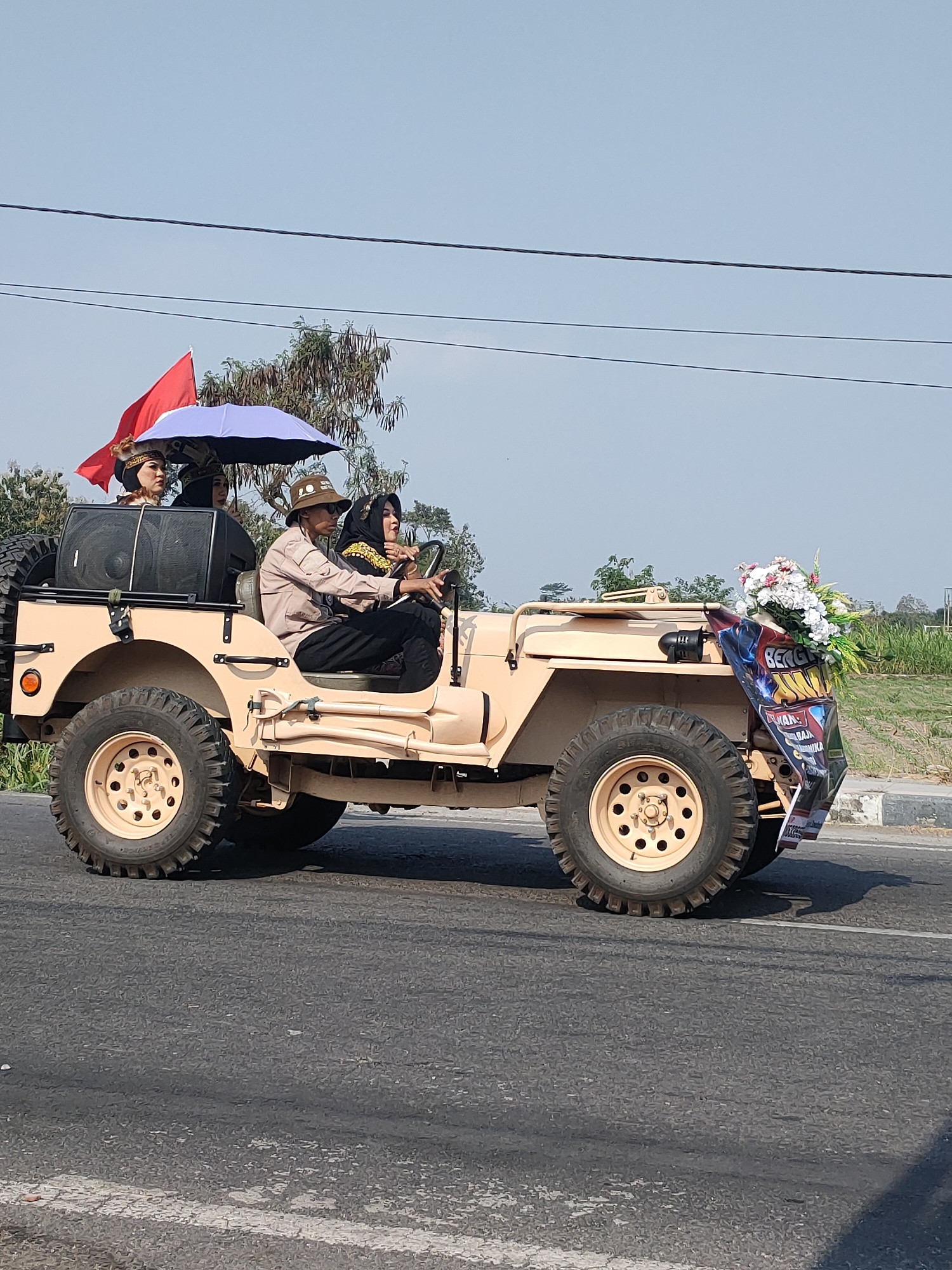 A beige, open-top jeep is driving on a paved road. Three people are in the jeep. A man wearing a hat and a light brown shirt is driving. A woman wearing a black hijab and a yellow, embellished top is in the passenger seat. Two women wearing headscarves and traditional attire are in the back seat, one holding a red flag and the other holding a blue umbrella. The jeep is decorated with a banner and flowers. The background shows trees and a clear sky. The scene is in the city of Kanigoro, Indonesia.