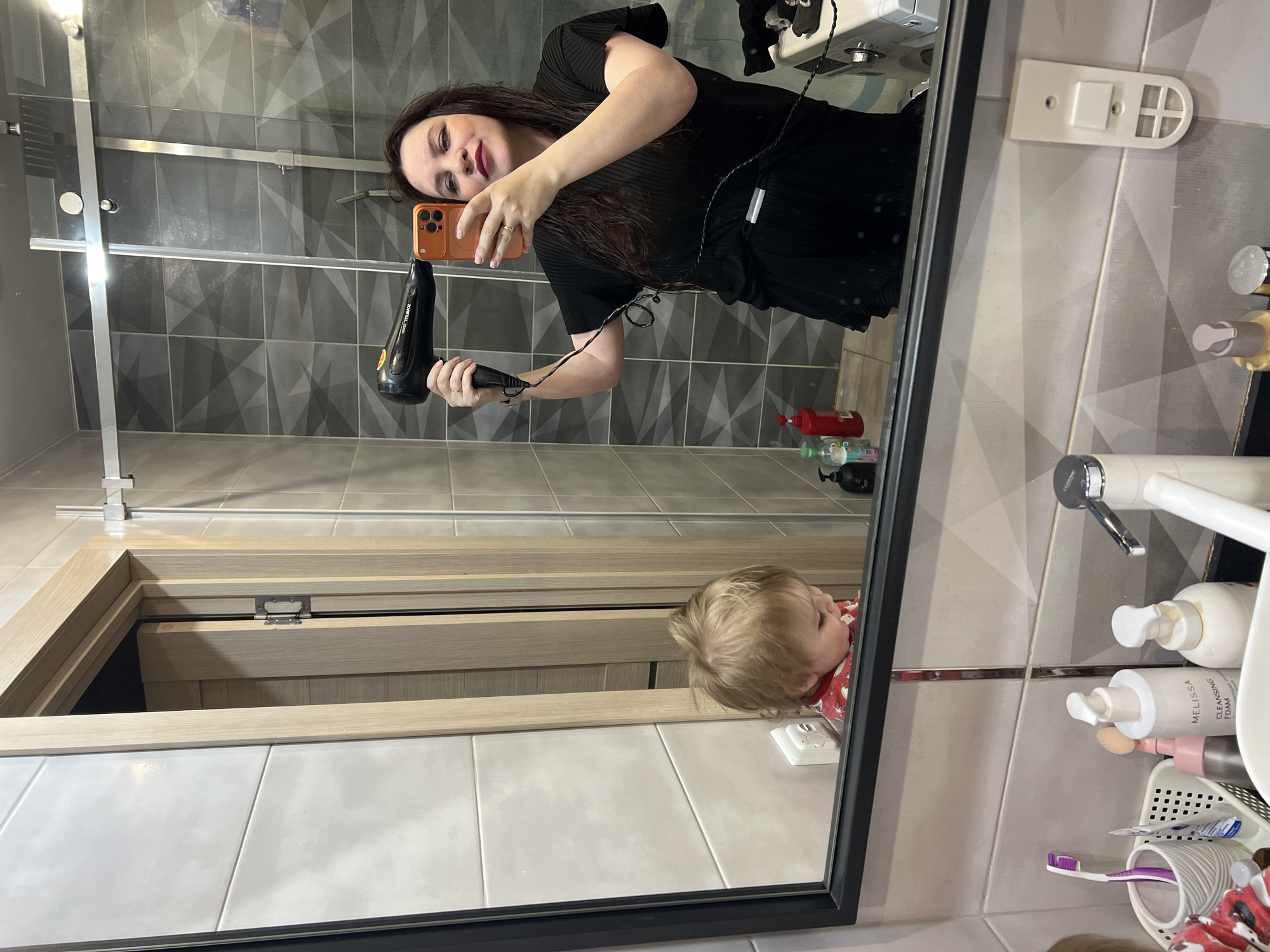 The image shows a woman in a bathroom, taking a selfie in a mirror. She is holding an orange phone in one hand and a black hair dryer in the other. She is wearing a black top and has dark hair. In the mirror's reflection, a young child with blonde hair can be seen. The bathroom has gray and white tiles. There is a shower, a sink with various toiletries, and a door in the background. The image may have been taken in the city of Noviy, Russia.