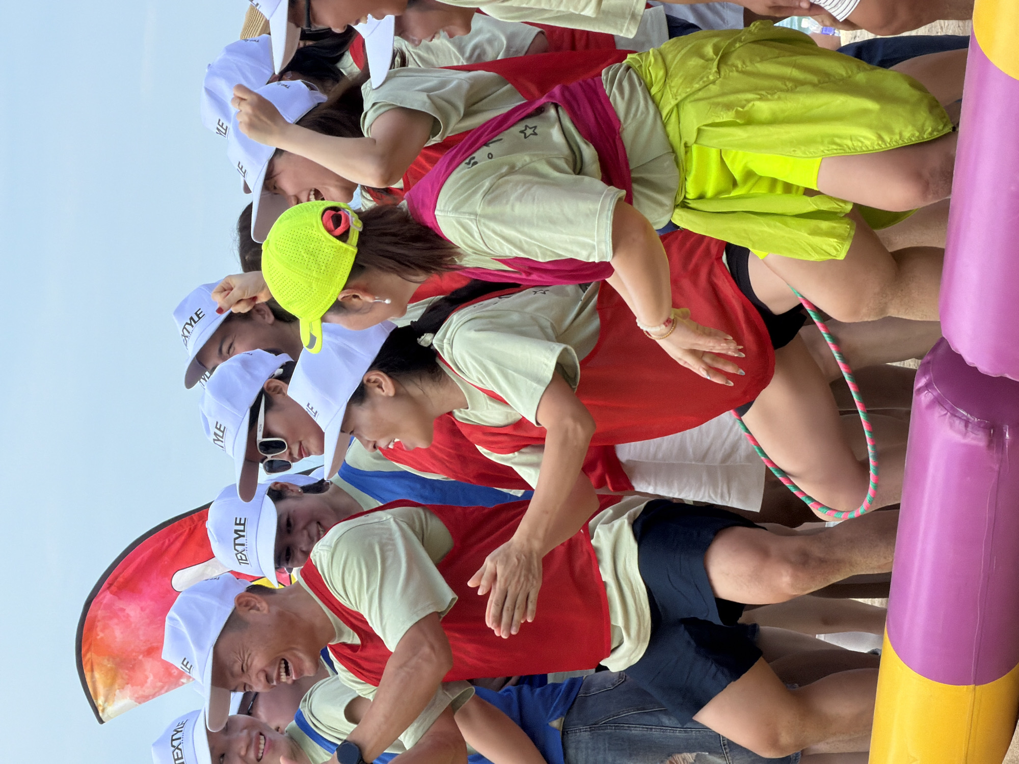 A group of adults in white caps with "TEXTYLE" printed on them are participating in an outdoor activity. The individuals are wearing light green t-shirts and red vests. Some are laughing and appear engaged in a game or competition. In the foreground, a colorful inflatable tube is visible. The background suggests an outdoor setting with a light-colored sky. The scene depicts a moment of group activity and enjoyment. The presence of the inflatable tube and the attire of the participants hint at a team-building event or recreational activity, possibly on a beach or open ground. The overall impression is one of lively participation and camaraderie.